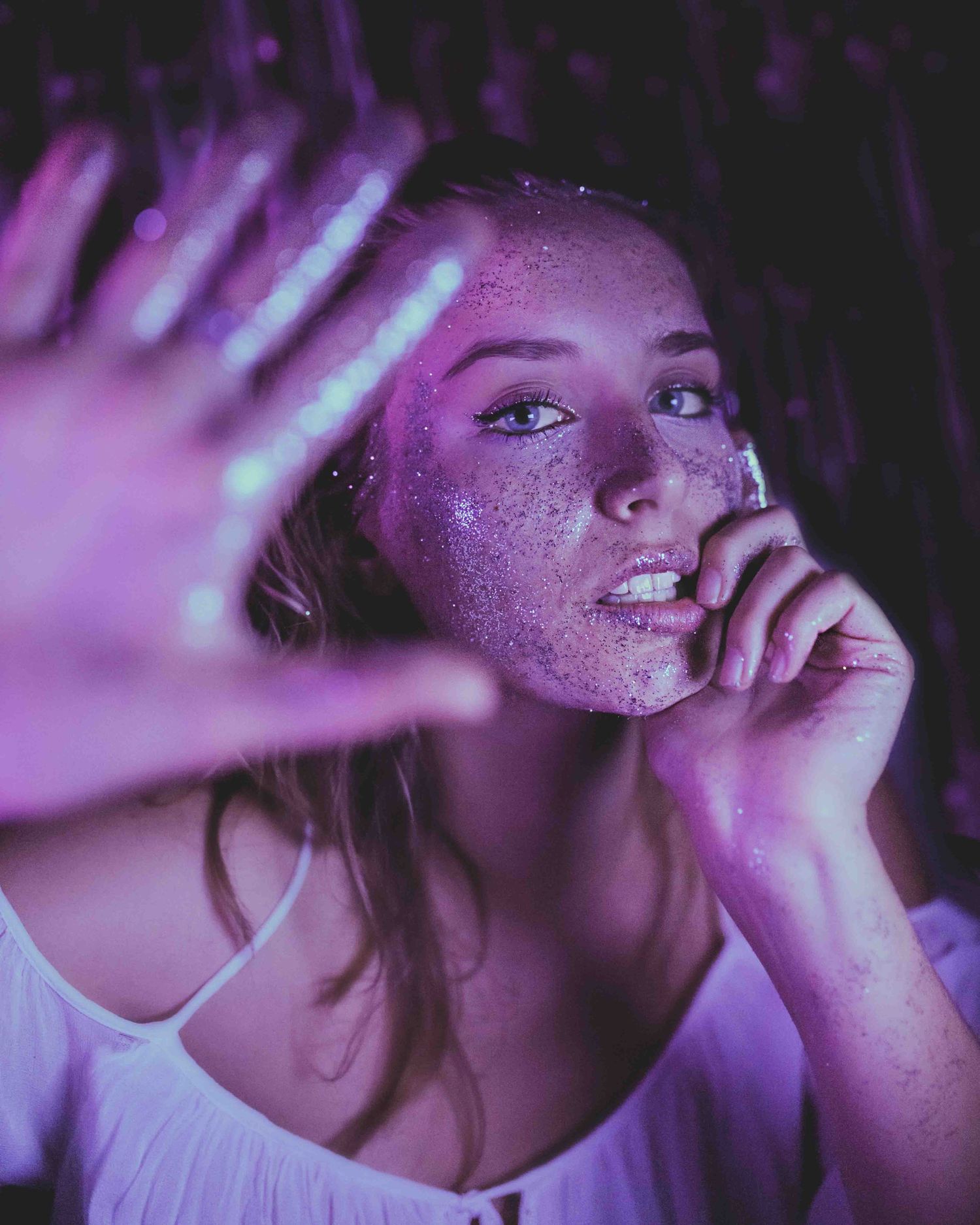 Artistic portrait with glitter makeup in purple lighting and a raised hand creating a dreamy ethereal effect.