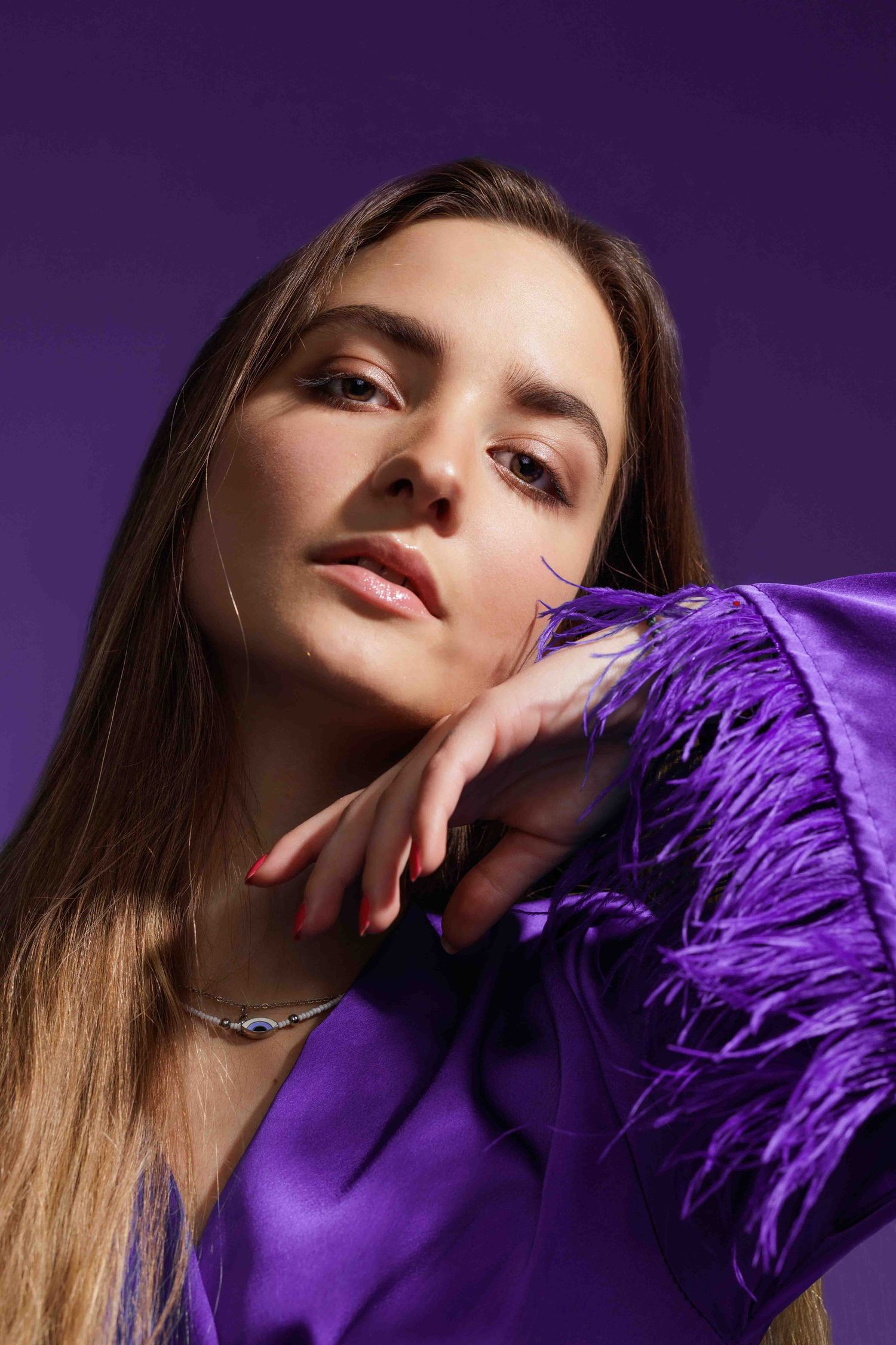 Fashion model wearing dark purple feathers poses dramatically against a purple-tinted backdrop.