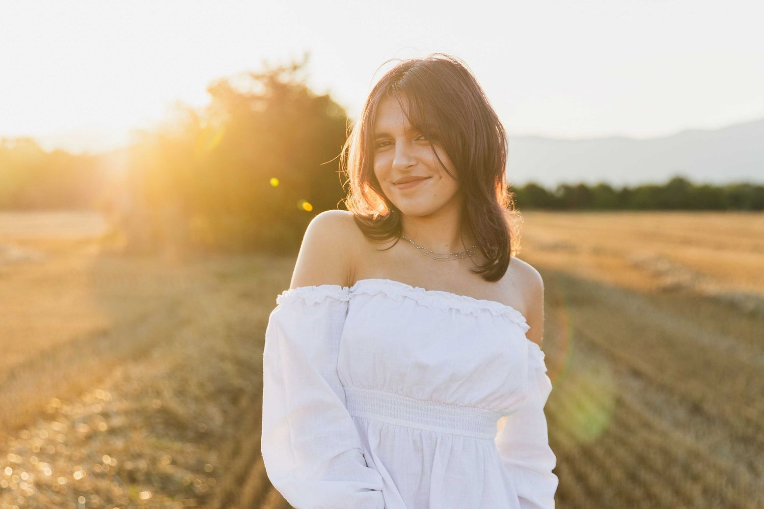 Person in white off-shoulder dress poses in golden sunlit field during sunset with lens flare and warm lighting.