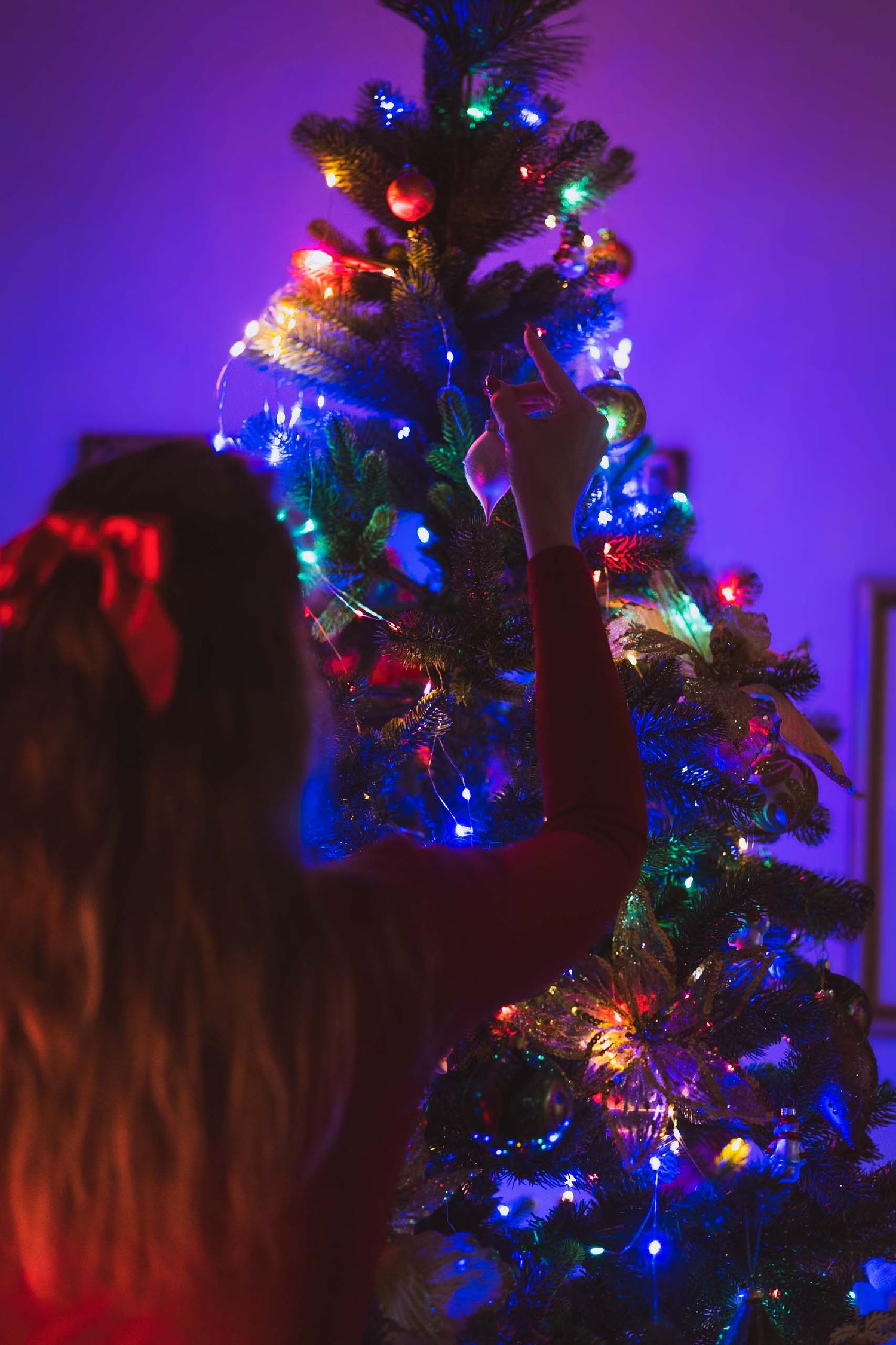 A silhouetted figure decorates a Christmas tree illuminated with vibrant blue and multicolored lights in a dark room.