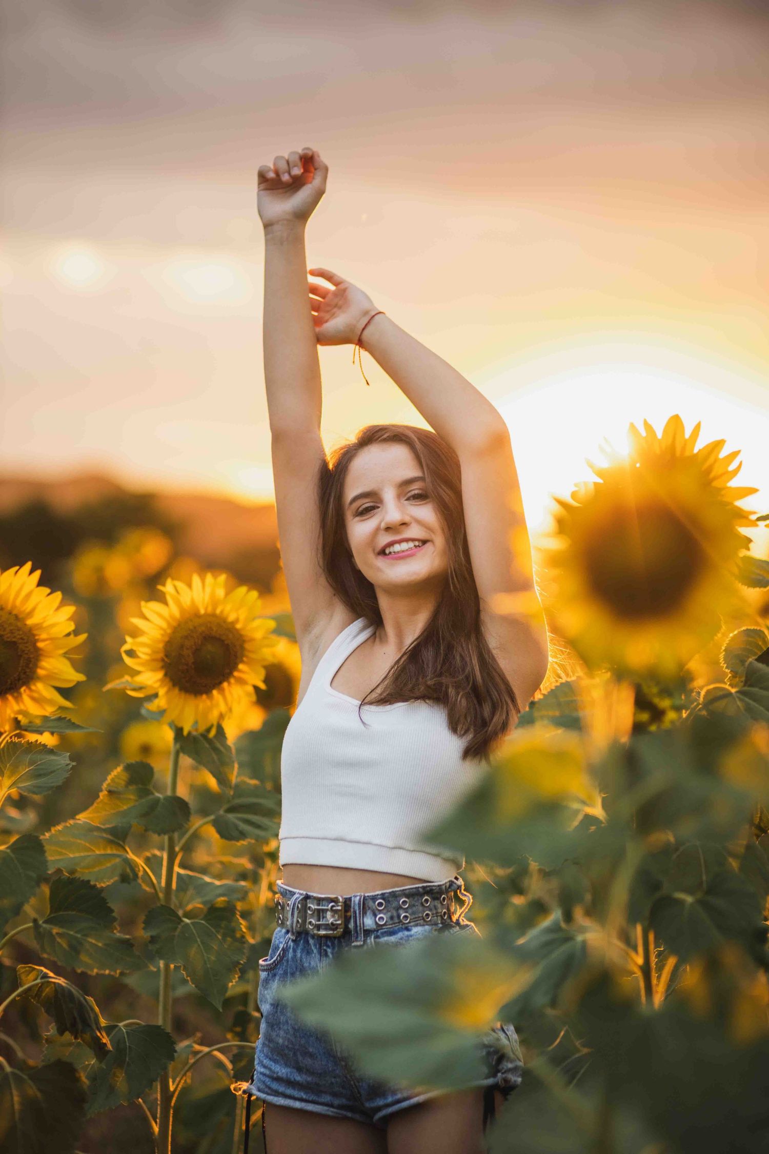 Figure in a sunflower field at golden sunset raising arms joyfully among bright yellow blooms and green leaves.