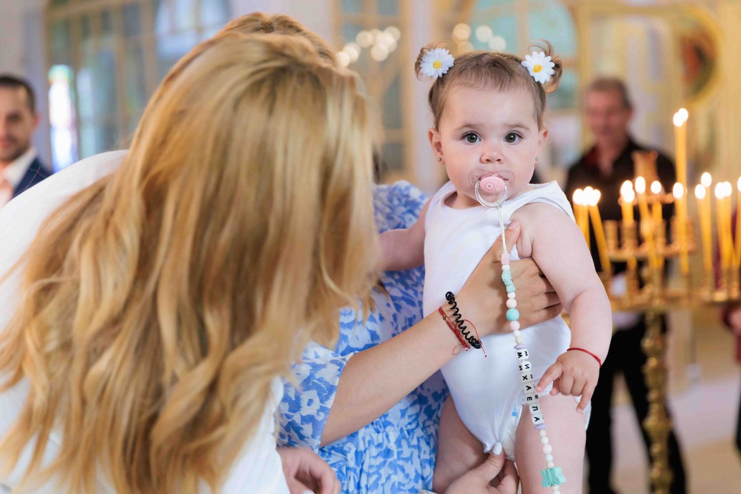 A small baby in white clothing is held during what appears to be a church ceremony with lit candles in the background.