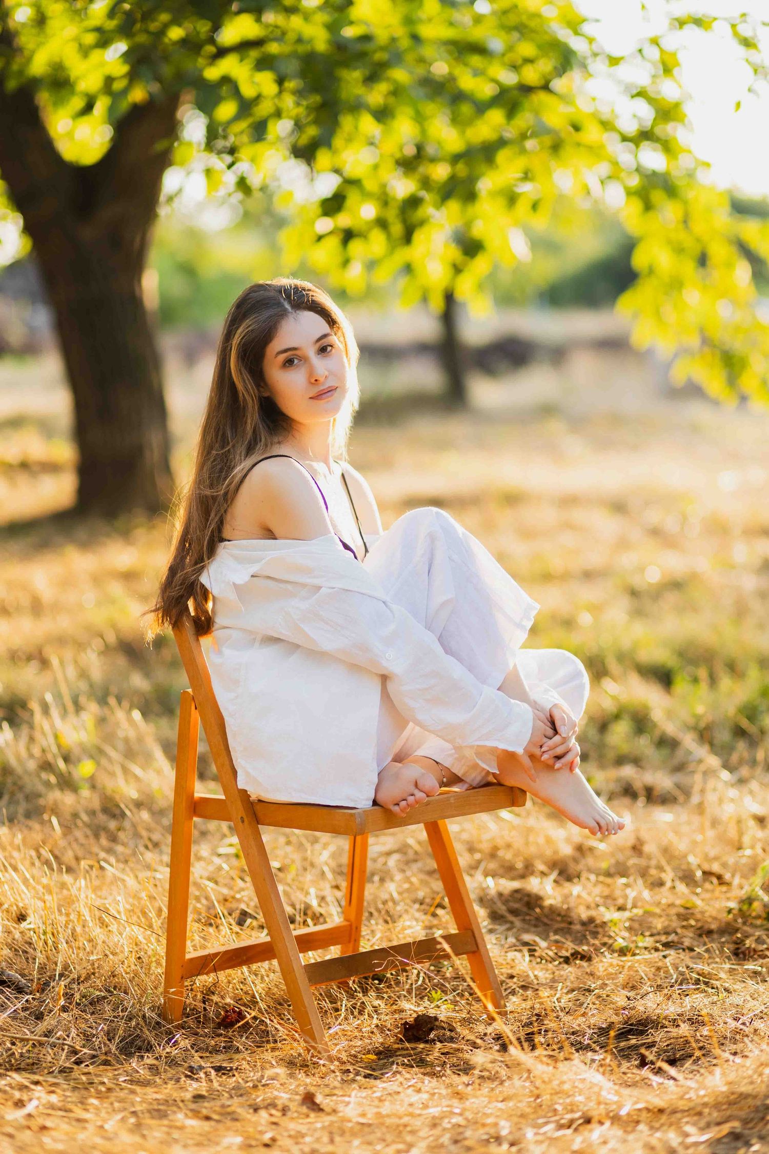 Portrait in a white outfit seated on wooden chair under autumn trees with warm sunlight filtering through leaves.