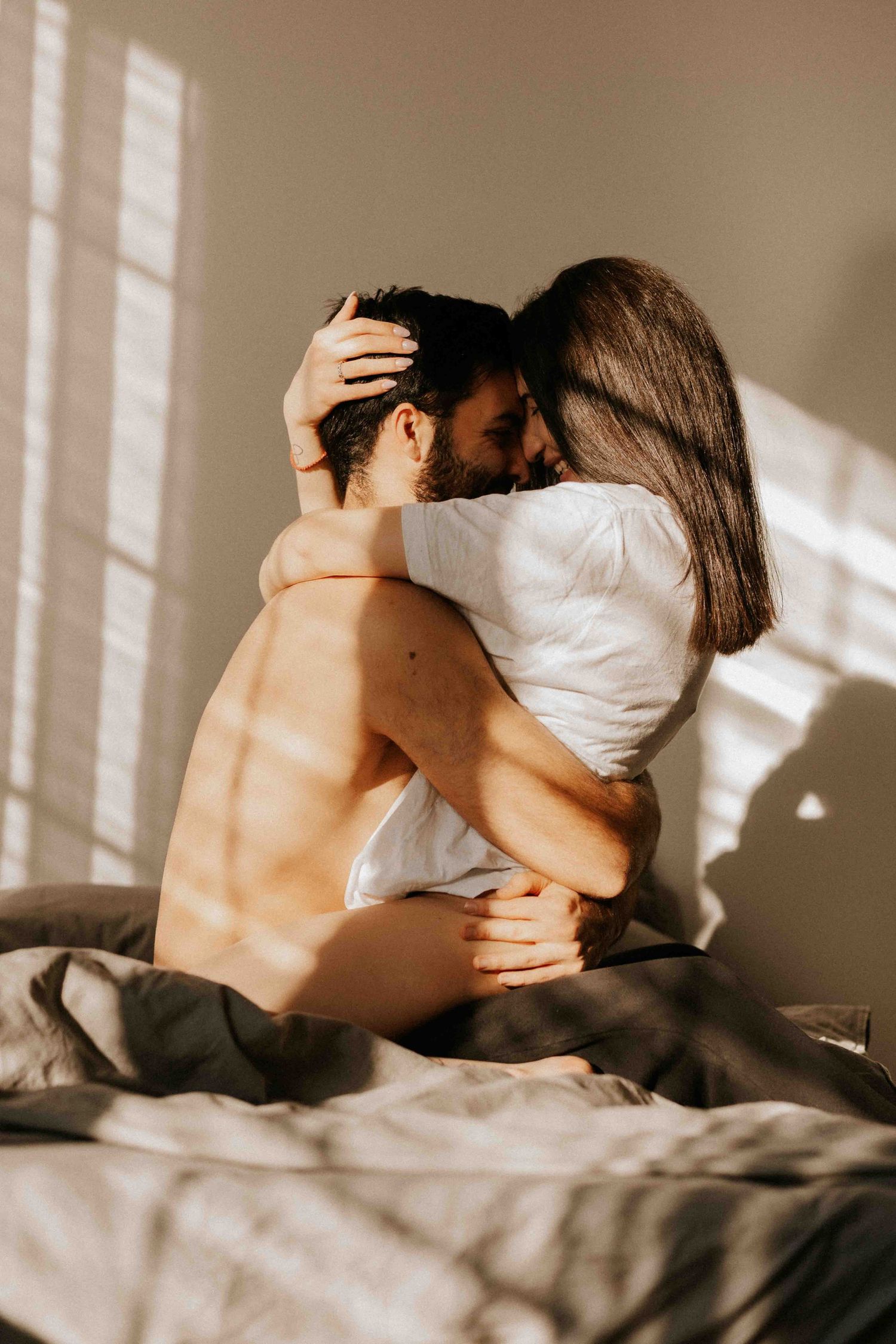 A romantic couple embraces intimately on a bed with sunlight streaming through window blinds creating shadows.