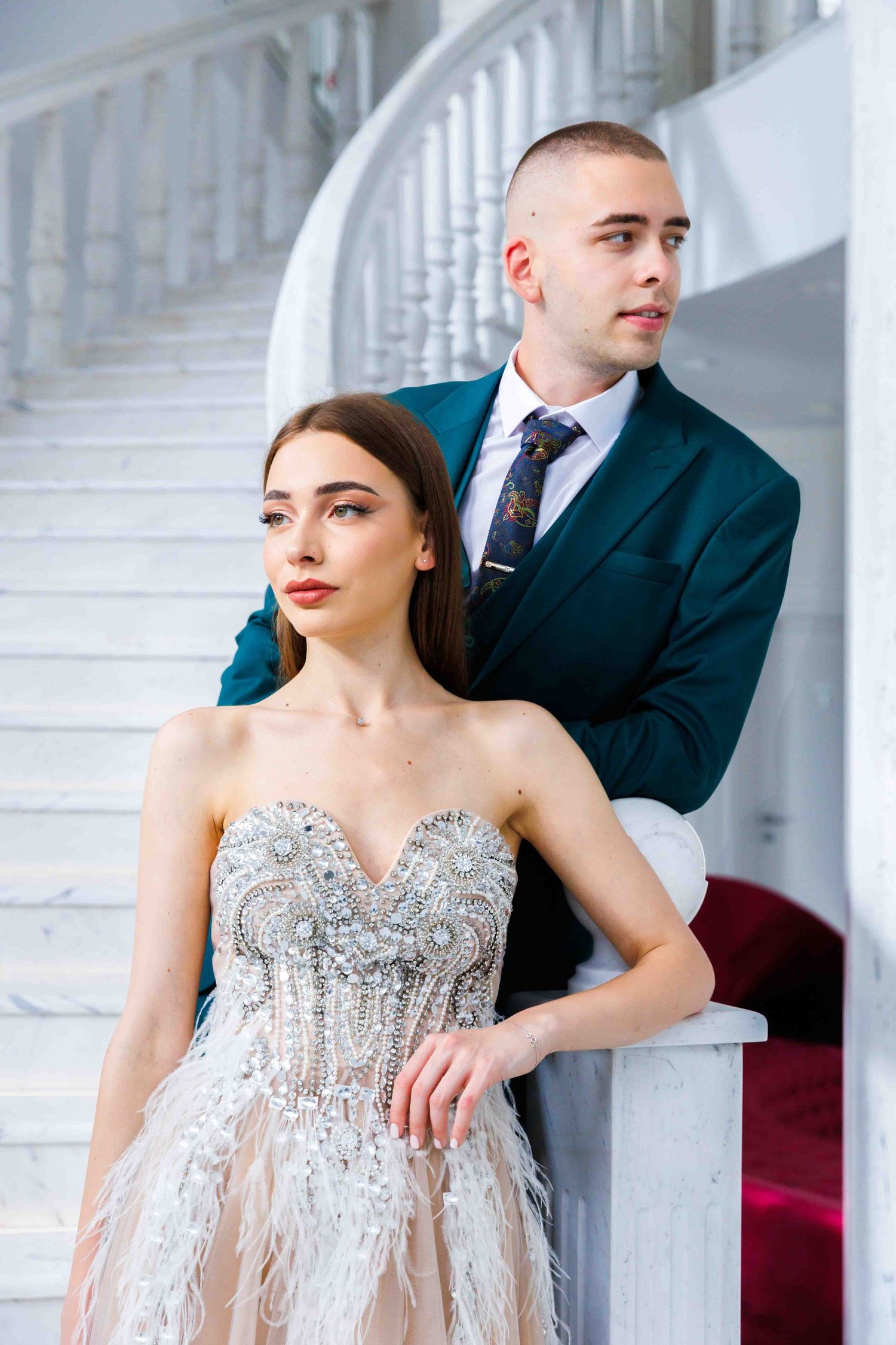 Glamorous couple in formal attire pose elegantly on white staircase in classic interior setting.