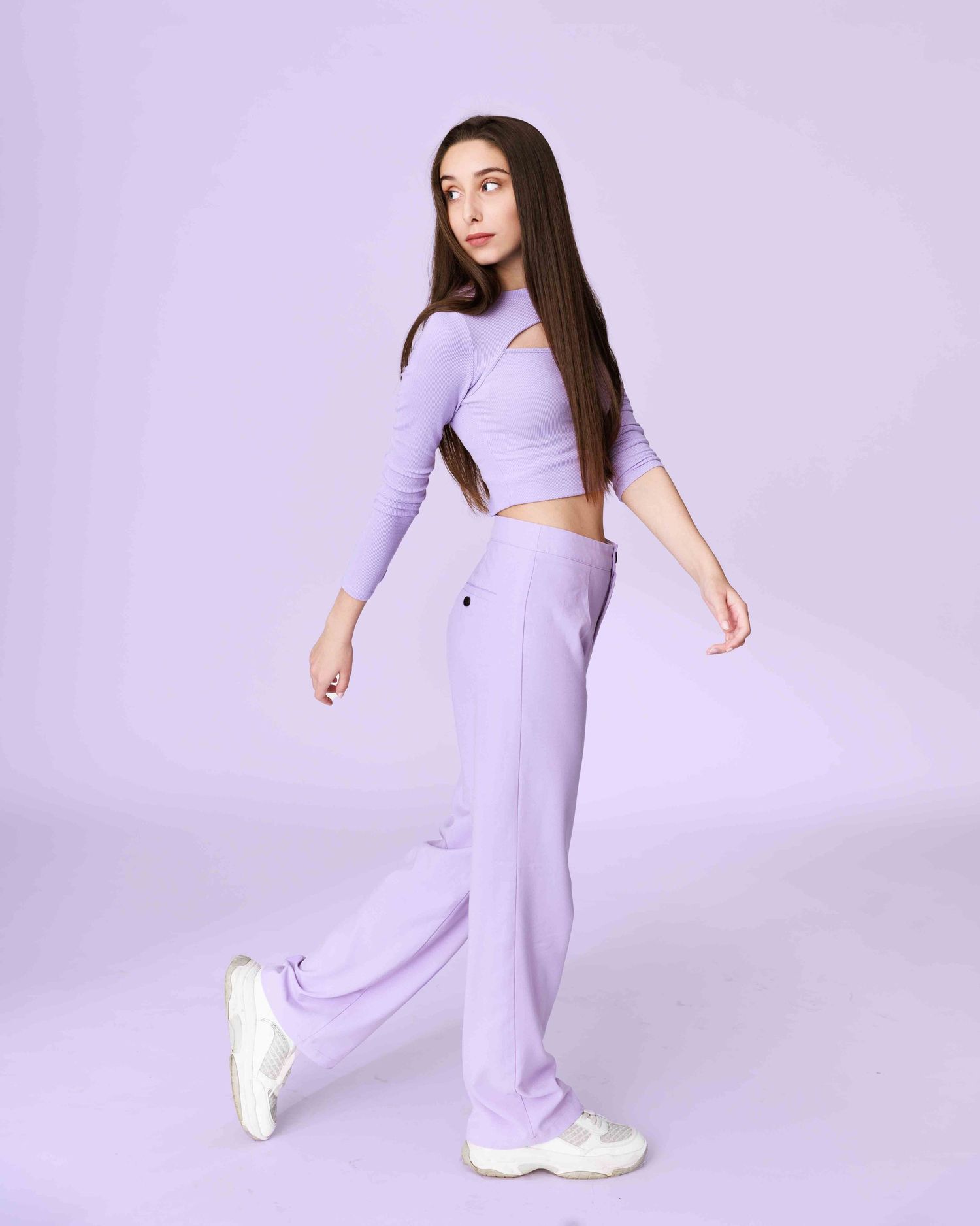 A model in lilac athleisure coordinates poses dynamically against a minimalist light purple studio backdrop.