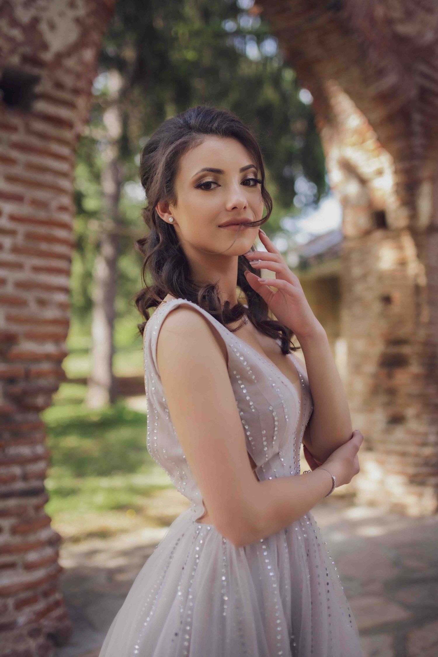 Woman in sparkly silver dress poses playfully against brick columns in outdoor garden setting.