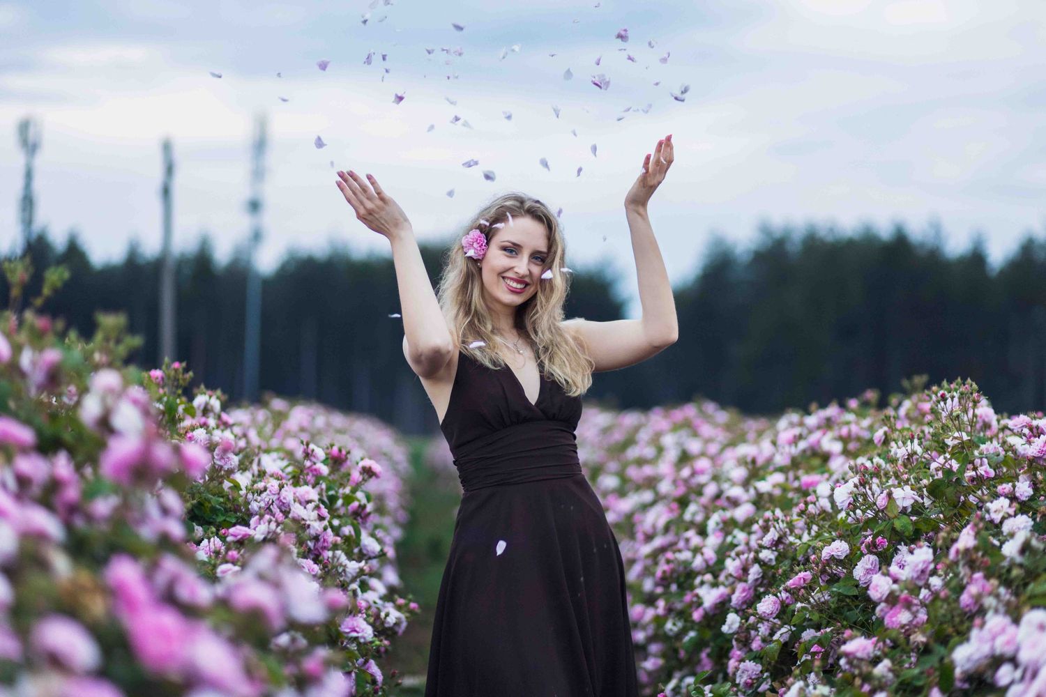 Person in a black dress celebrates joyfully amid a field of vibrant pink flowers on a cloudy day.