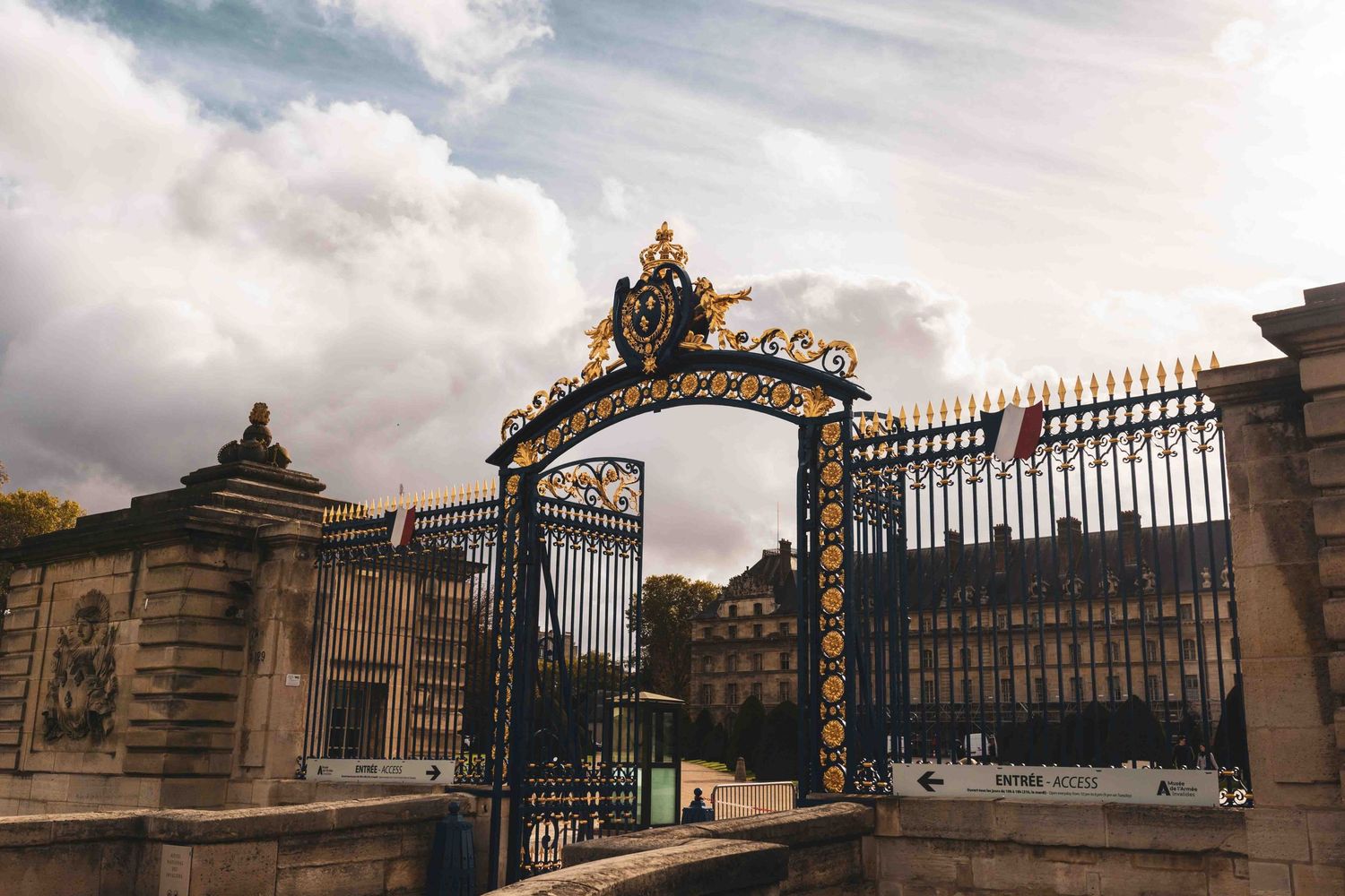 Ornate wrought iron gates and stone pillars guard the entrance to a historic estate on a cloudy day.