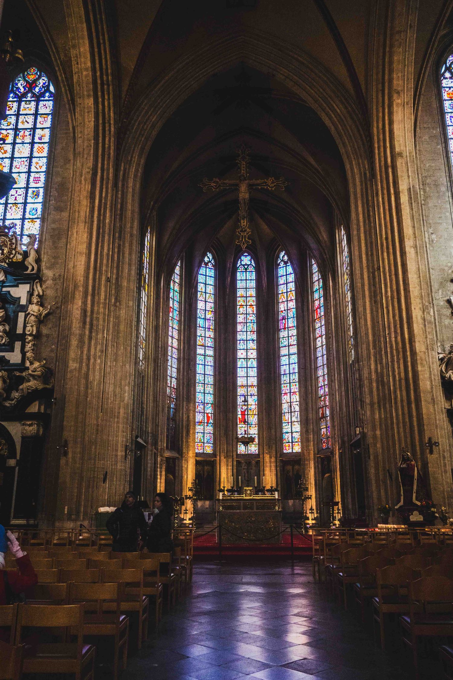 Interior view of a Gothic cathedral with tall stained glass windows, vaulted ceilings, and wooden pews leading to the altar.