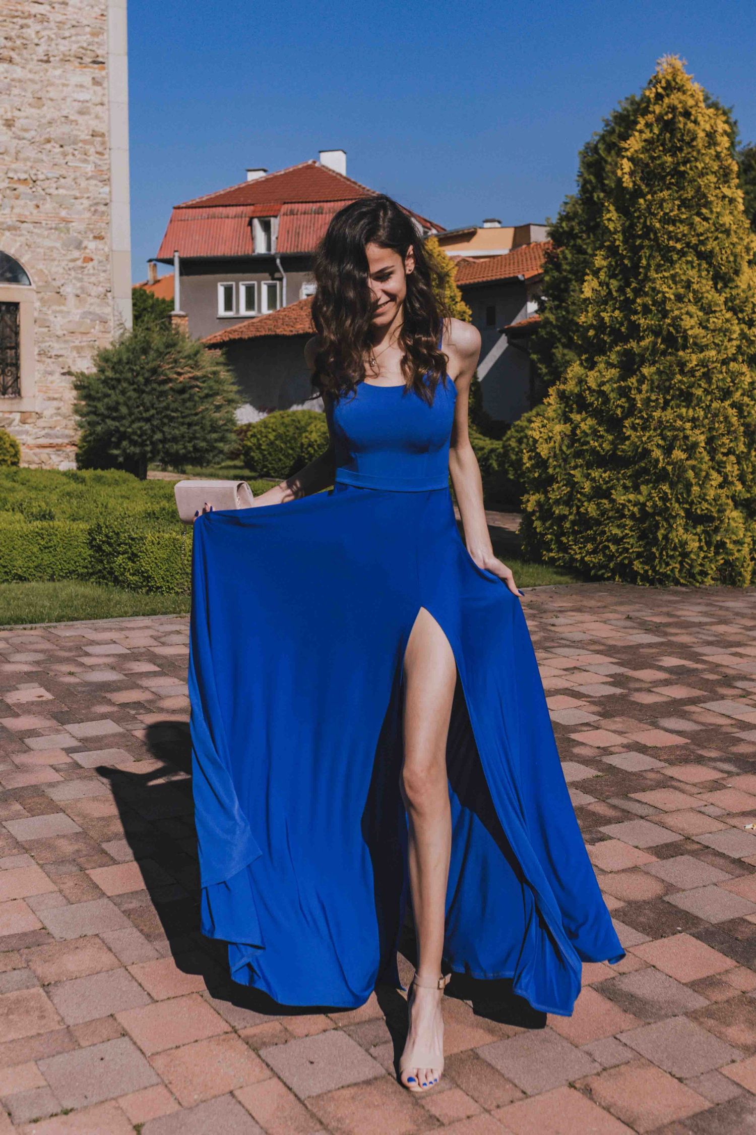 A striking royal blue evening gown with a thigh slit flows elegantly against a brick patio backdrop on a sunny day.