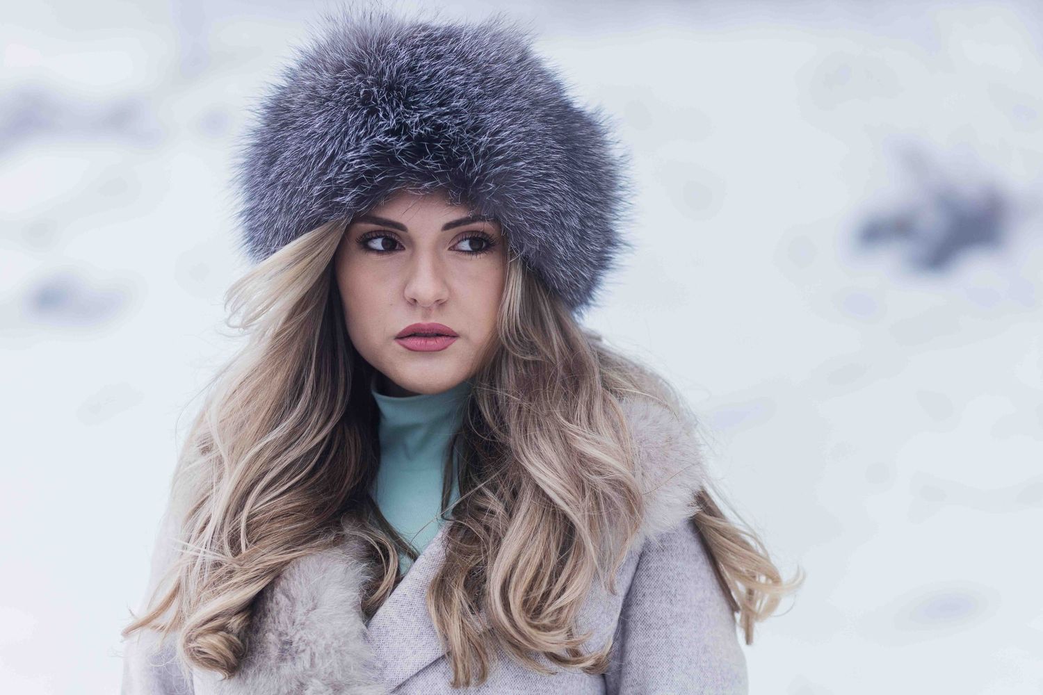 A winter fashion portrait featuring a plush fur hat and light blue turtleneck against a snowy backdrop.