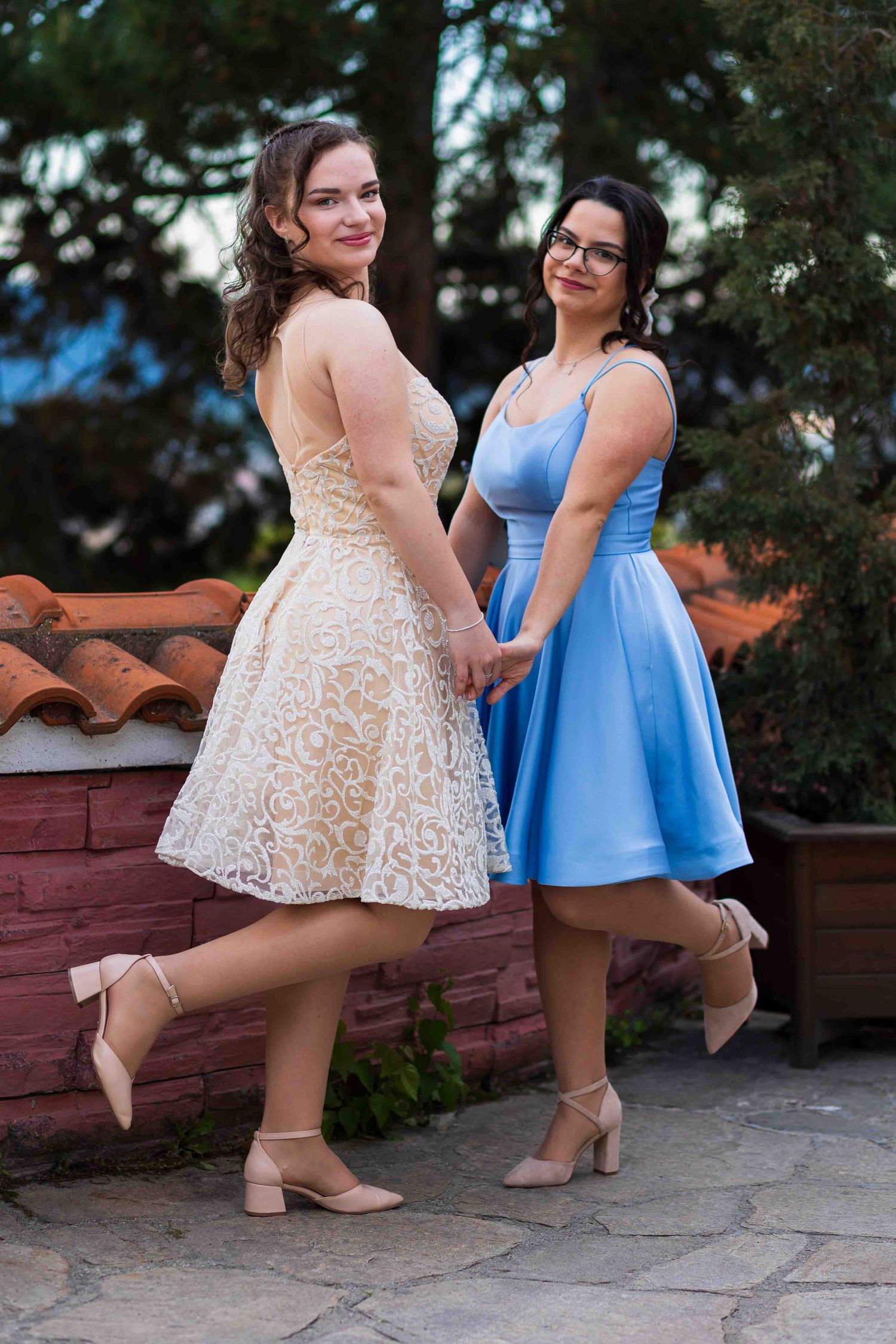 Two women pose in short party dresses - one in cream lace and one in light blue - while wearing matching nude heels.