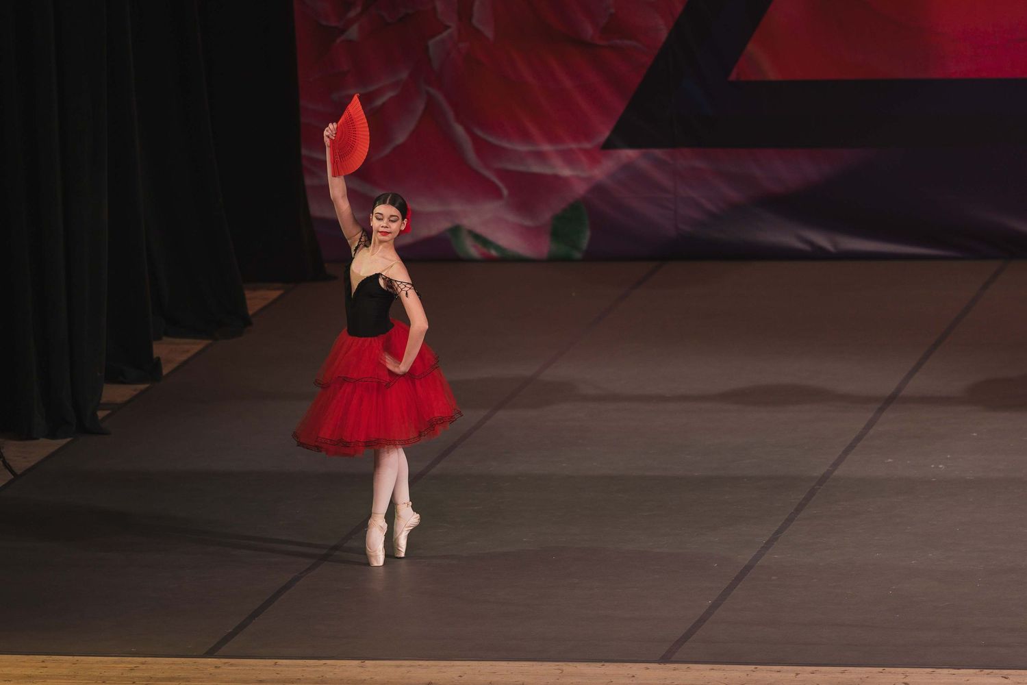 Ballet dancer performs on stage in a red tutu dress, striking an elegant pose during a recital performance.