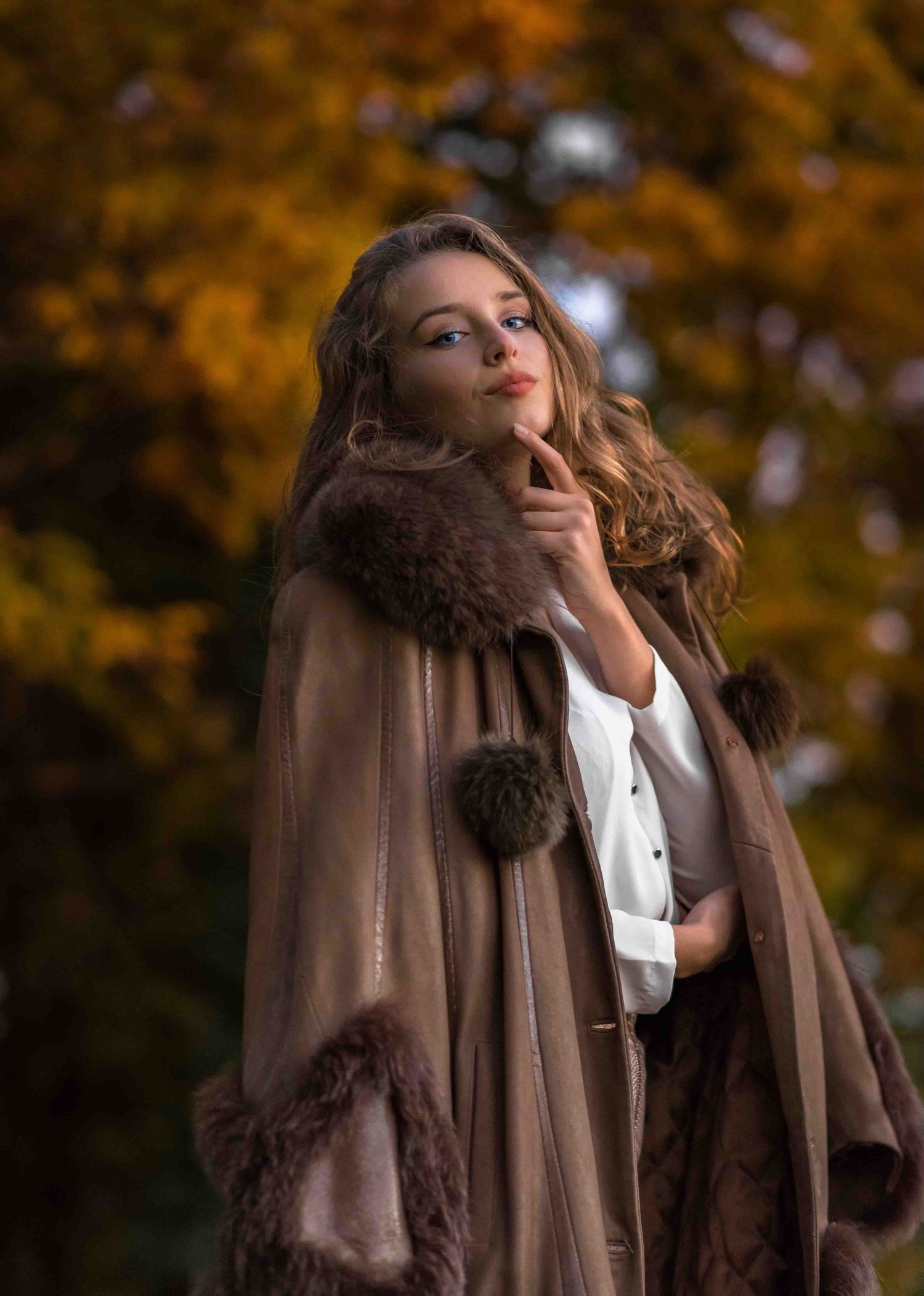 A model in a luxurious brown fur-trimmed cape poses thoughtfully against an autumn background with golden foliage.