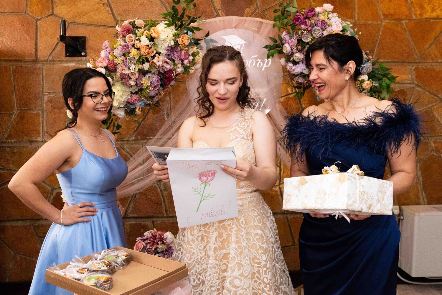Three elegantly dressed guests look at presents together under a floral archway at a formal celebration event.