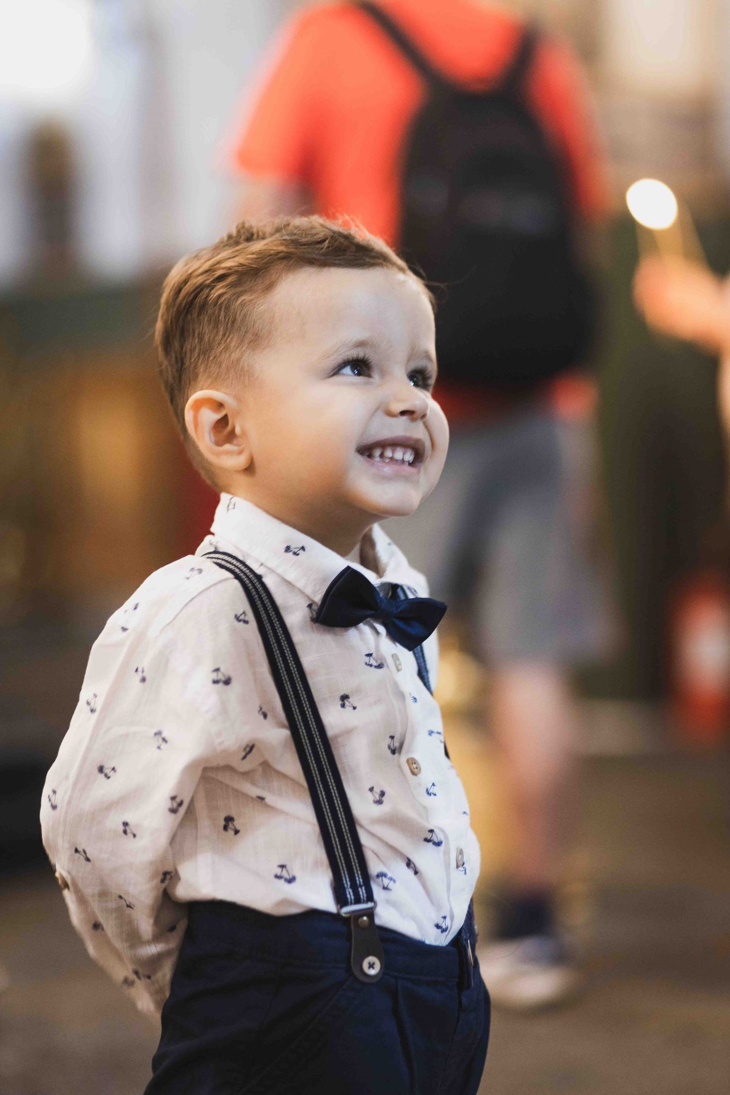 A young child in formal attire with suspenders and bow tie smiles while looking up at something off camera.