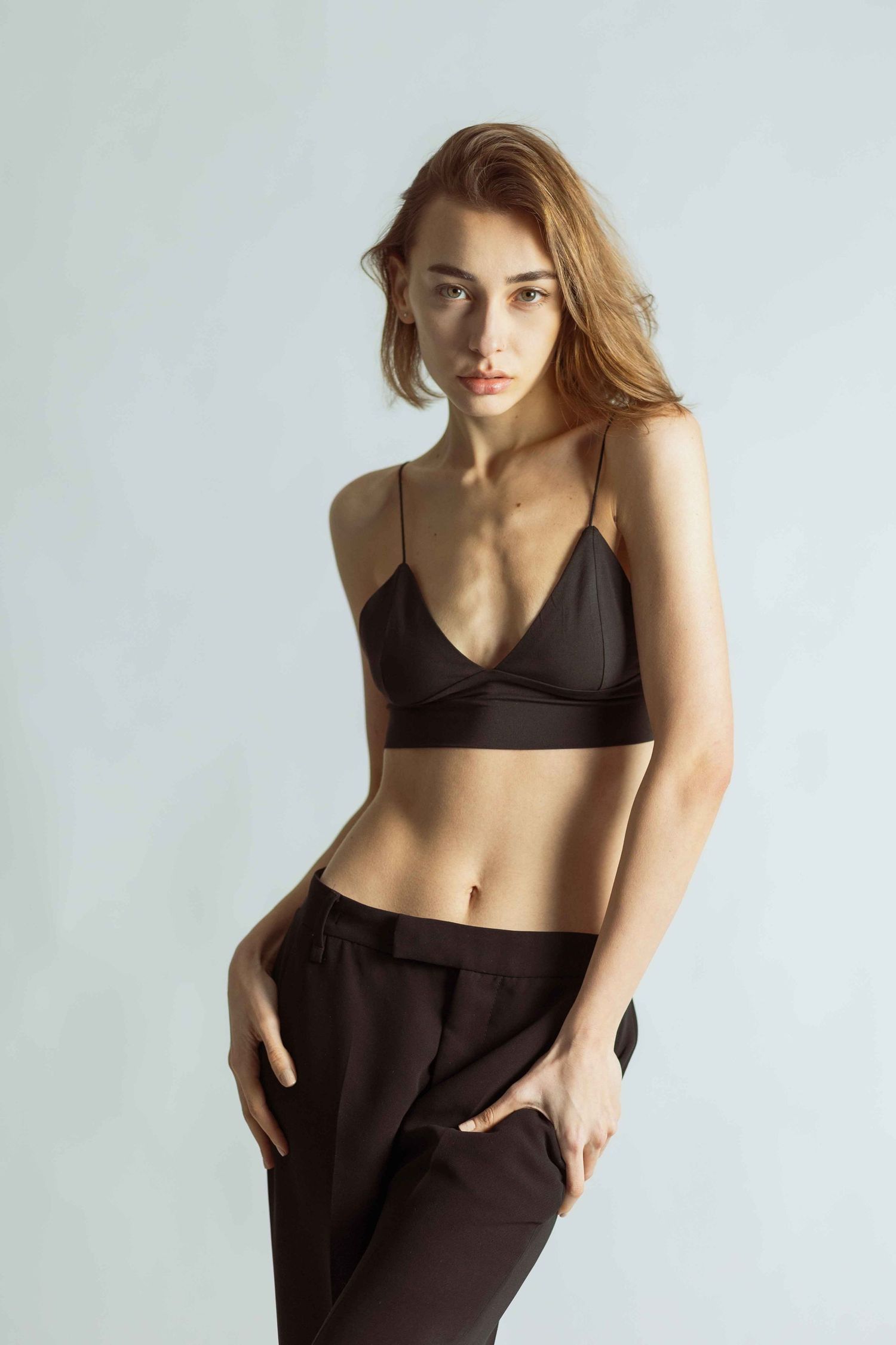 Fashion model wearing black sports bra and trousers poses against light background in minimalist studio setting.