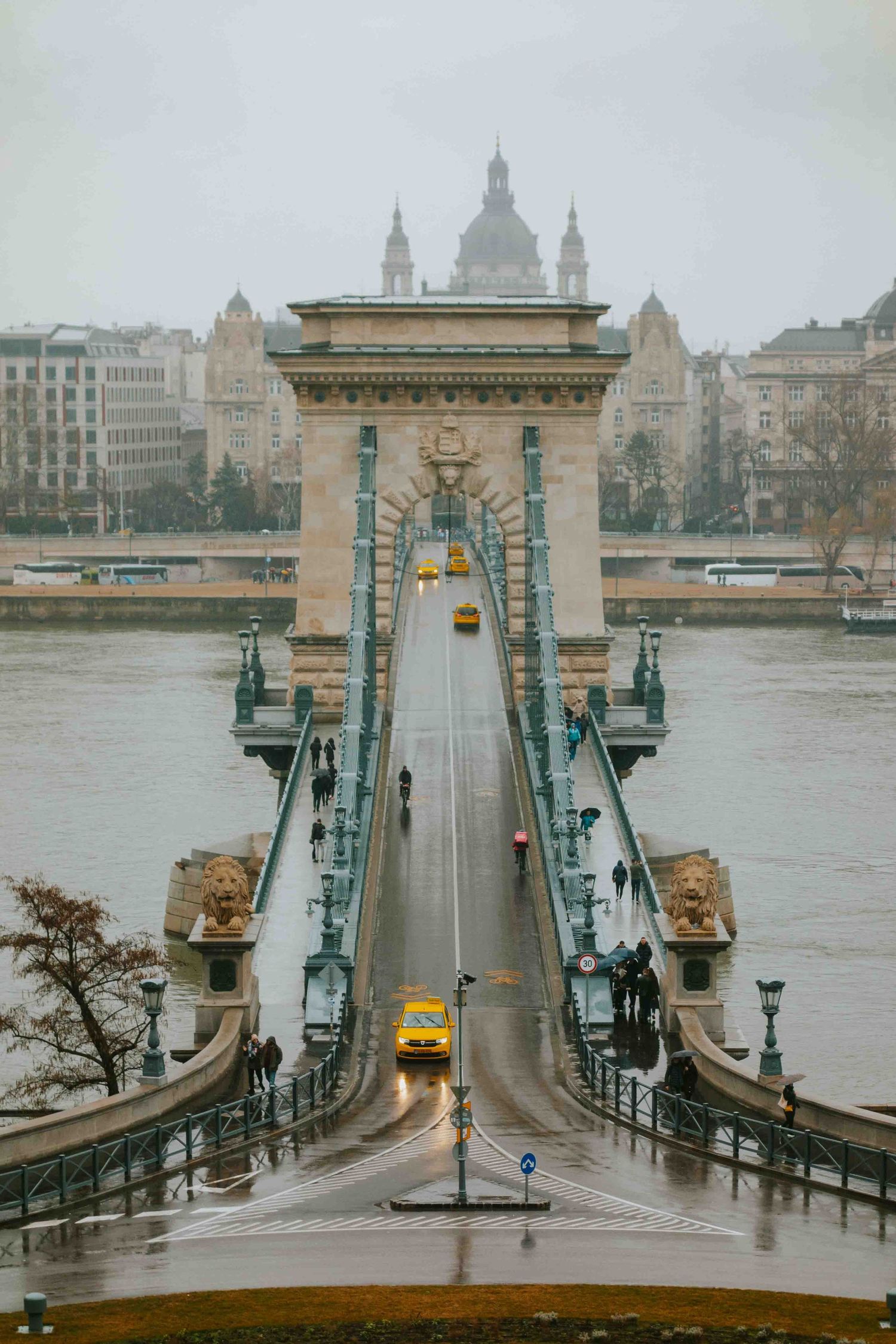 Yellow taxis cross the iconic Chain Bridge in Budapest on a rainy day with St. Stephen's Basilica dome in background.