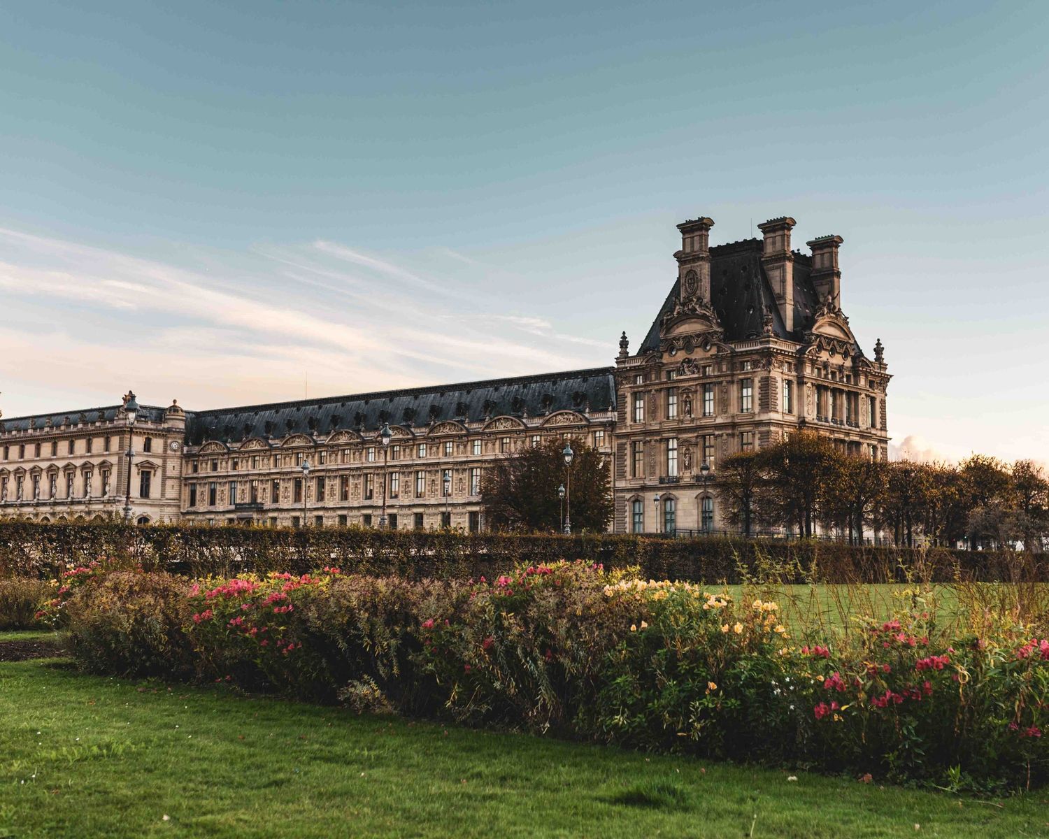 The historic Louvre Palace in Paris stands majestically against a blue sky with colorful gardens in the foreground.