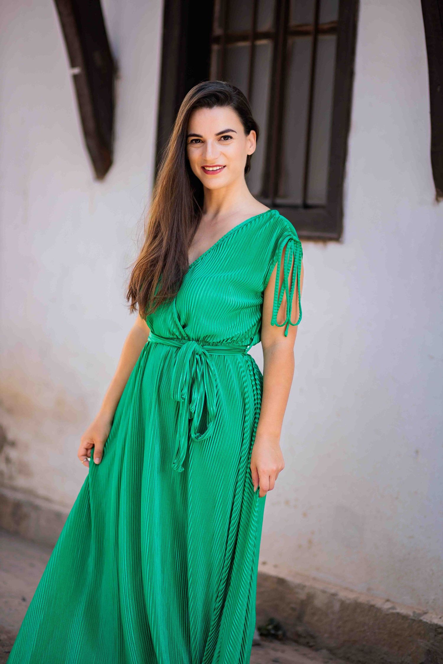 A woman in a flowing emerald green maxi dress with tie details stands against a white wall with dark trim.