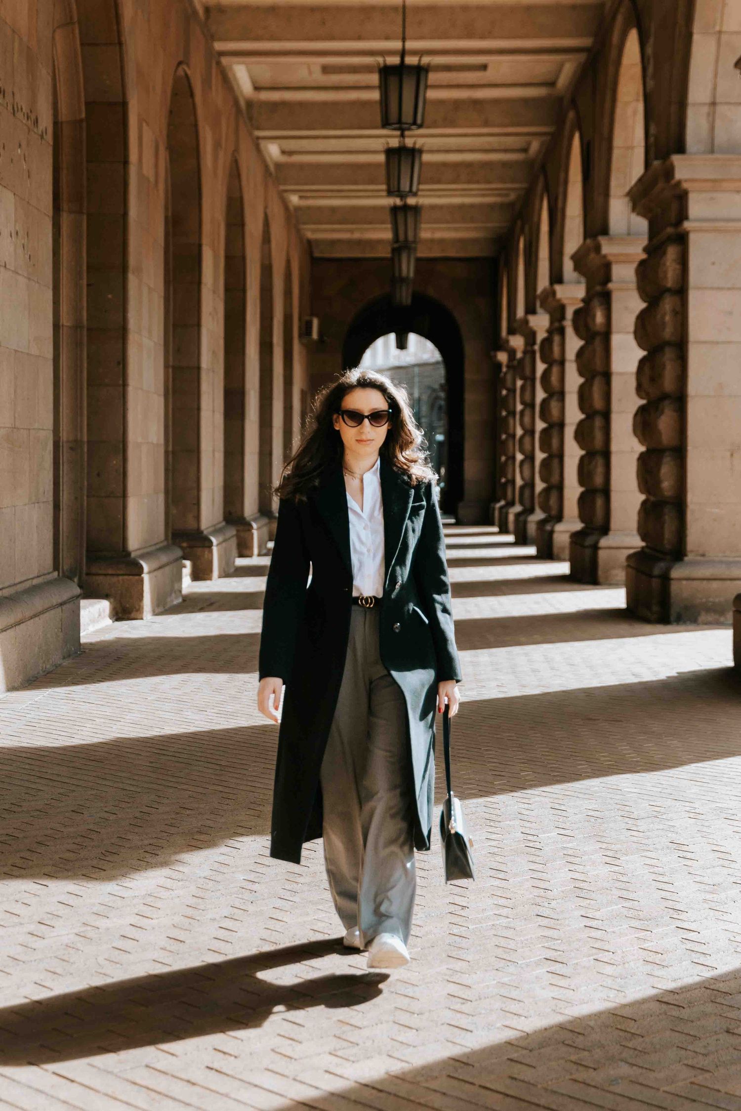 A figure in a black blazer and denim walks through a colonnade hallway with dramatic lighting and stone architecture.