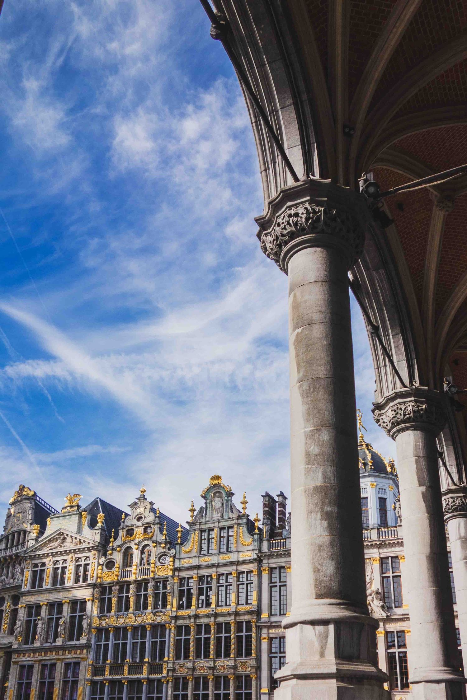 Ornate Gothic columns frame historic Belgian guild houses with decorative facades against a bright blue sky in Brussels Grand Place.