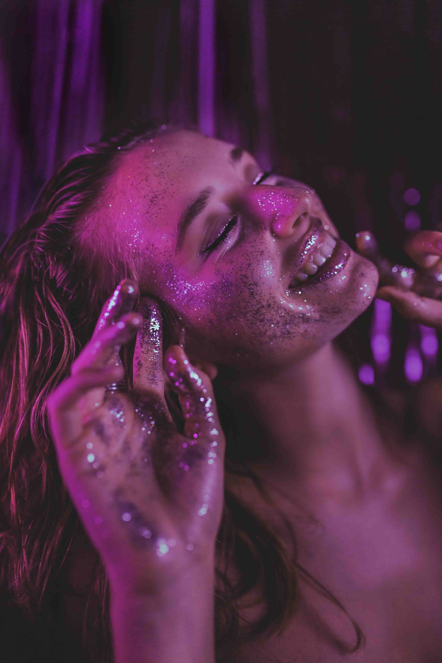 Artistic portrait bathed in purple light shows glittery skin and closed eyes in an ethereal dreamy pose.