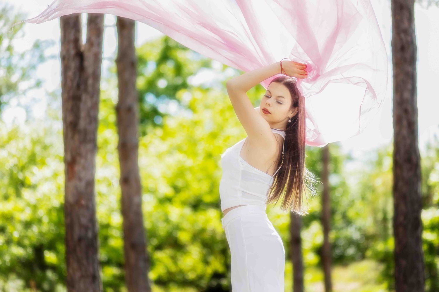 A graceful figure in a white dress twirls with flowing pink fabric in a sunlit forest setting.