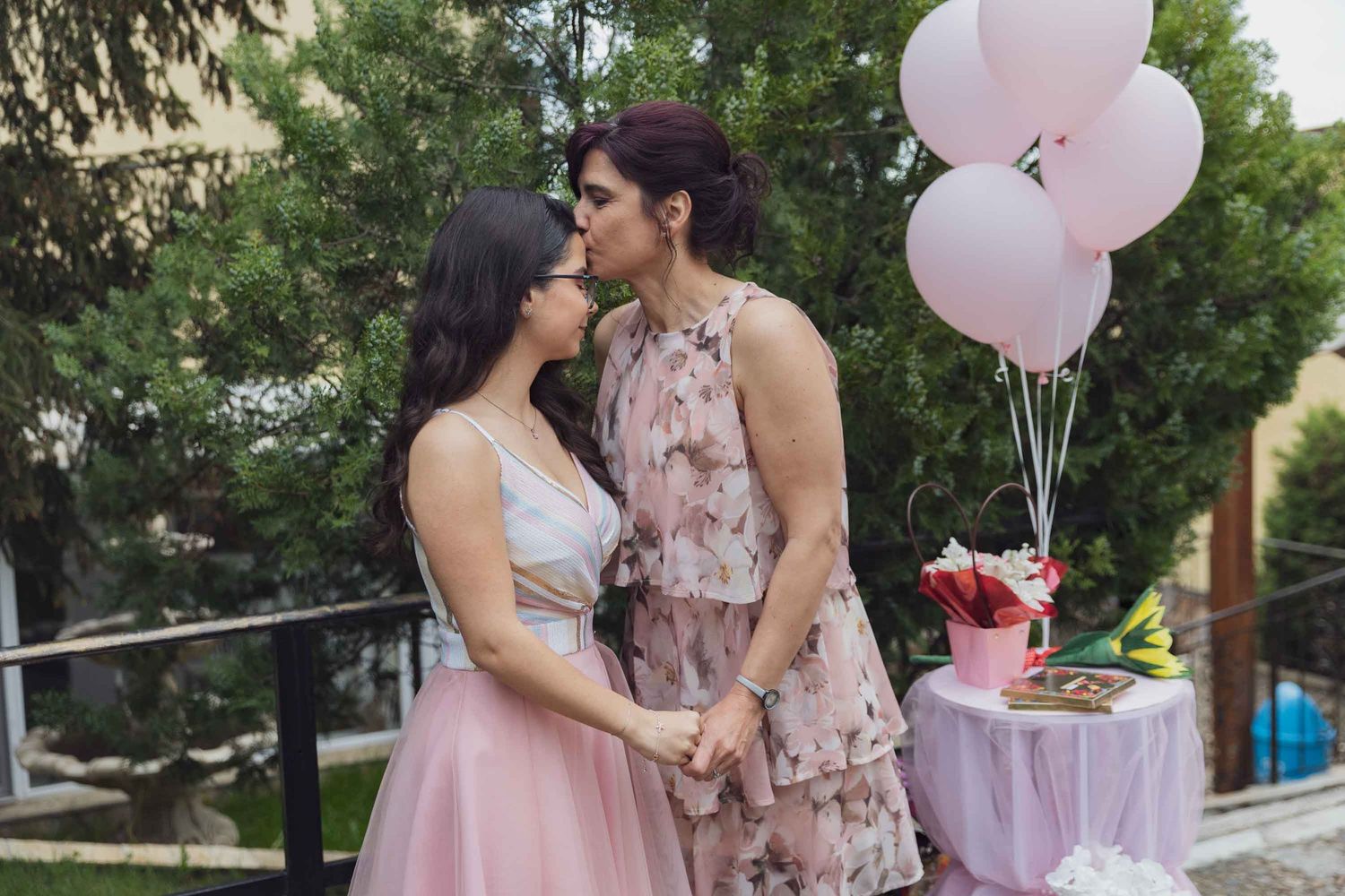 A romantic outdoor couple in pink dresses share a tender moment with pink balloons and a decorative table setting.