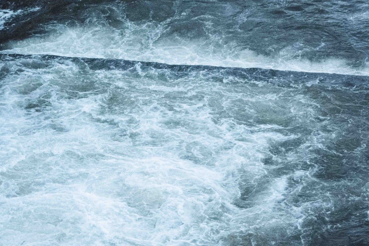 Turbulent blue-gray ocean water churns and swirls with white foam creating a dramatic pattern of waves.