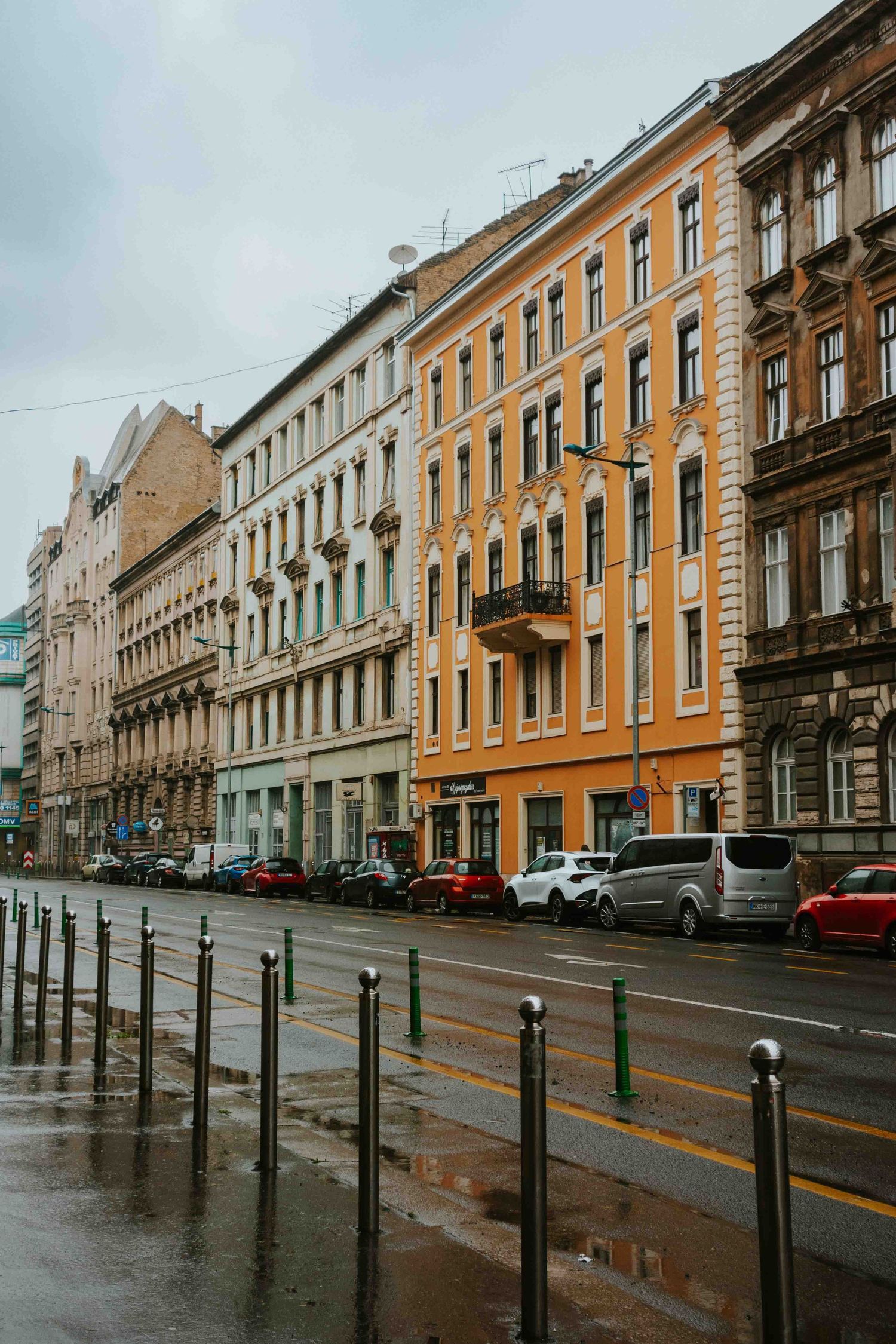 Moody urban street with historic colorful apartment buildings and parked cars along metal railings in rainy weather.