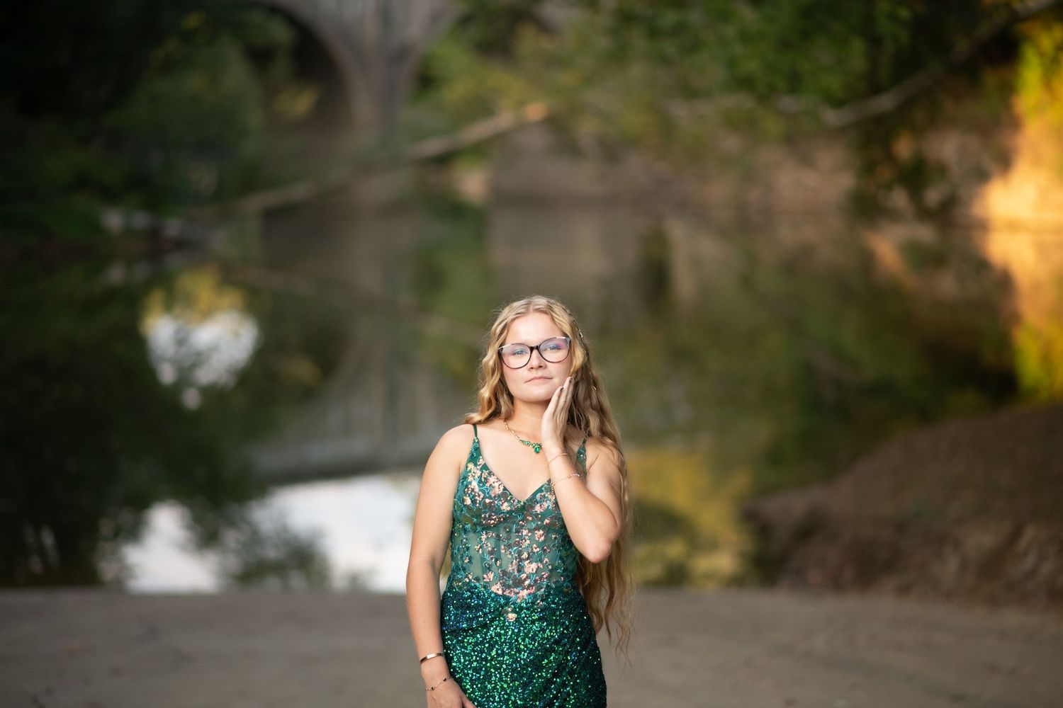 Young woman in green sequin dress with glasses poses near a stone bridge by a river in soft natural light.