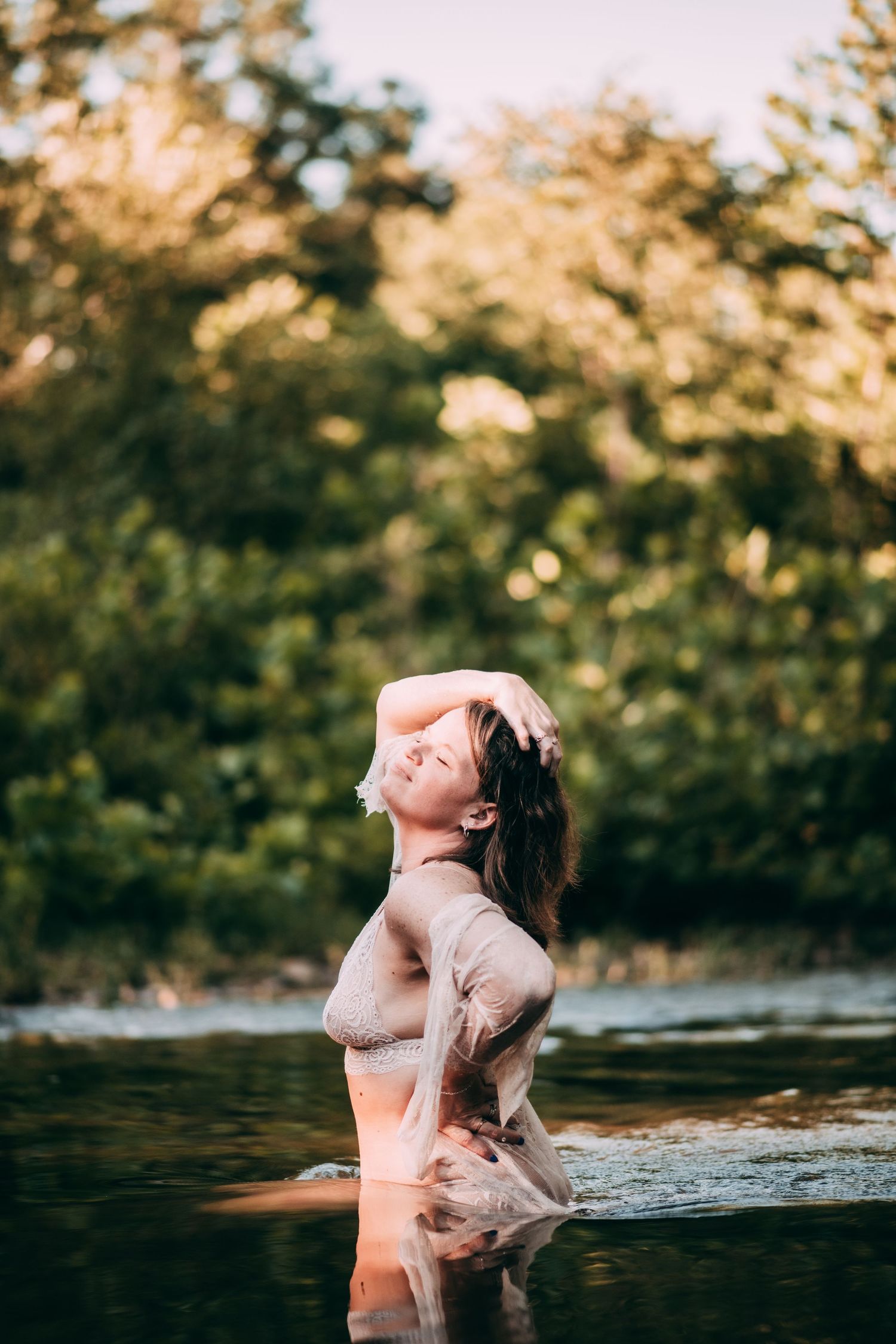 Woman standing in a river with wet hair, wearing sheer clothing, surrounded by lush green trees at golden hour.