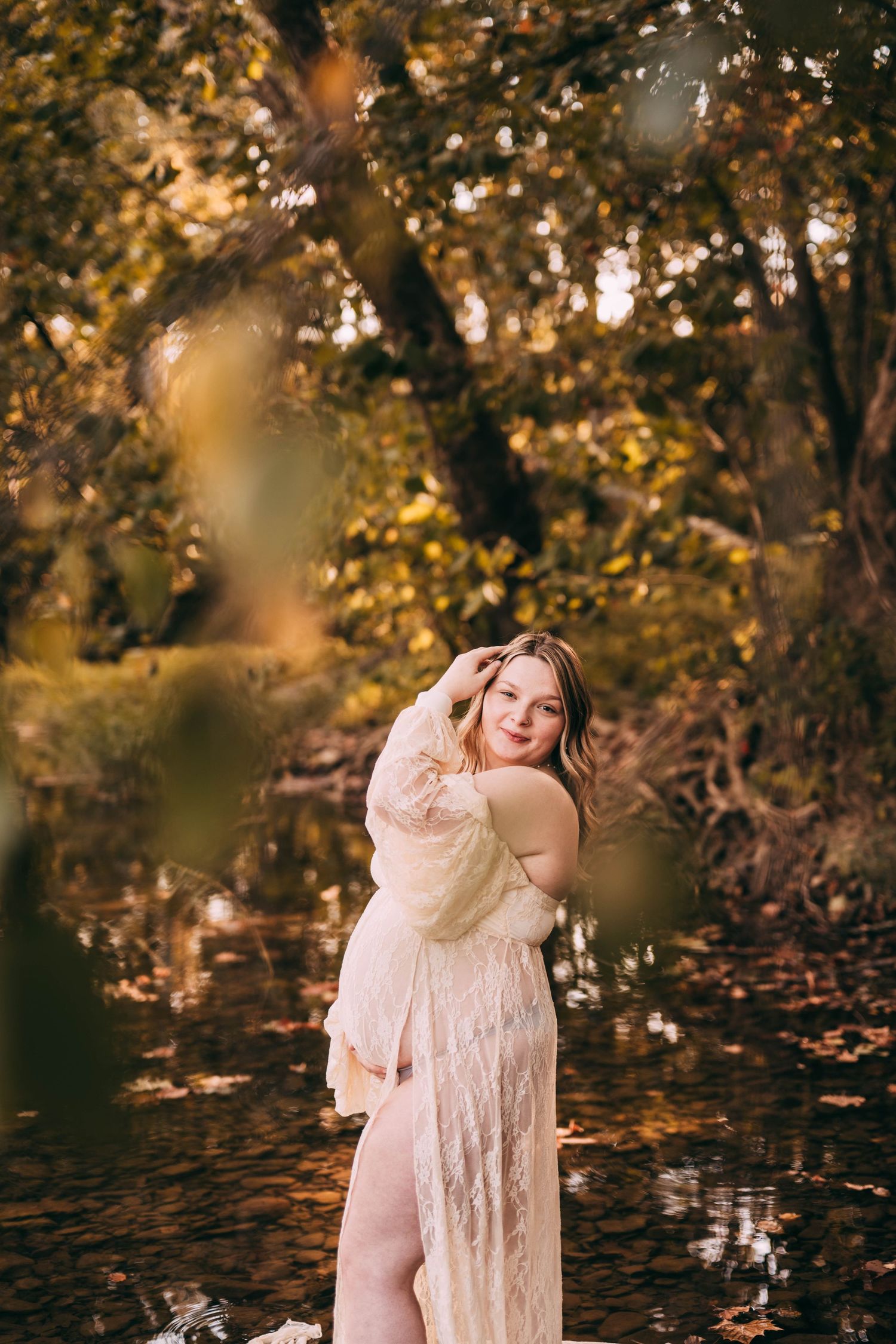 A person in a flowing lace dress poses gracefully by a stream in an autumn forest with golden sunlight filtering through trees.