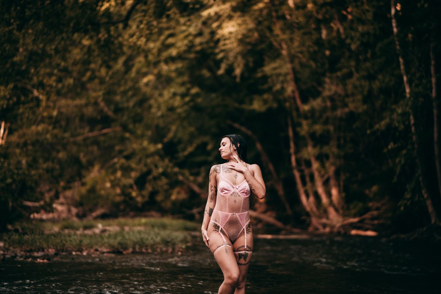 Woman in pink lace bodysuit standing in a forest stream surrounded by lush green trees. creek session, creek boudoir, Vanceburg Ky
