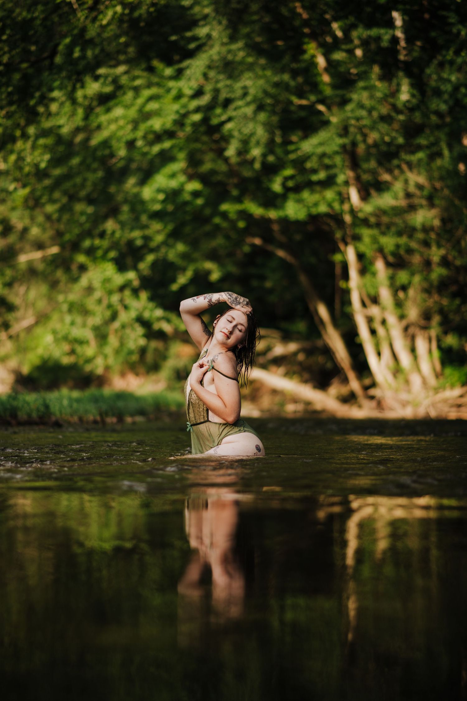 A woman stands waist-deep in a calm forest river, posing gracefully with lush green trees reflected in the water.