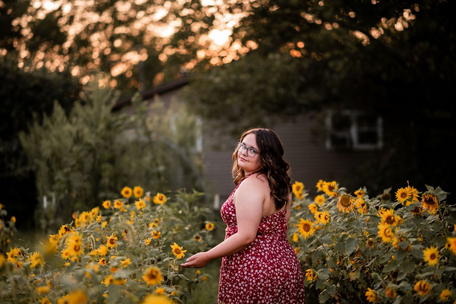 Woman in red floral dress standing among sunflowers at golden hour in a lush garden.