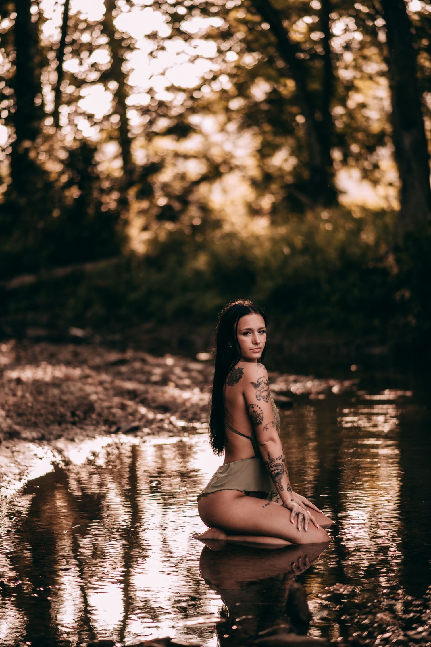Tattooed woman in bikini kneeling in a shallow forest creek, surrounded by golden bokeh trees.
