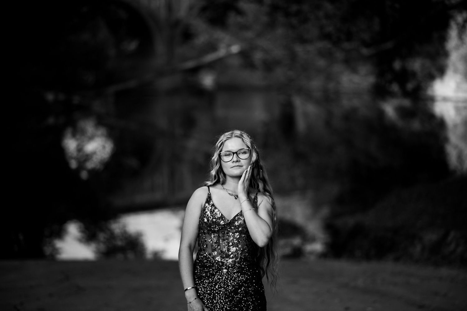 Black and white portrait of a young woman in a sequined dress wearing glasses, standing outdoors among trees.