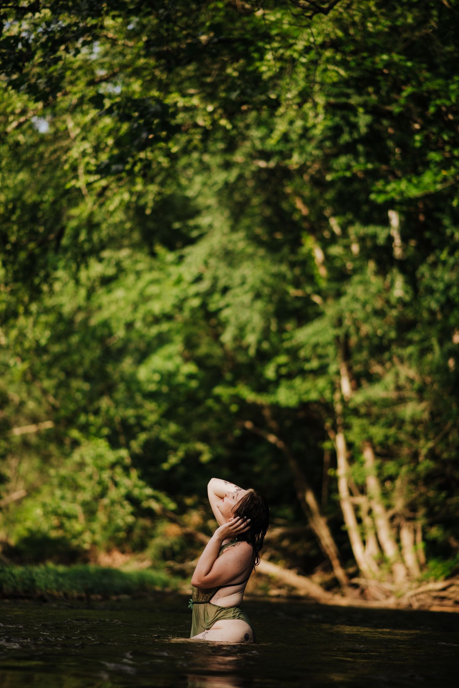 Woman standing in a forest river surrounded by lush green trees, head tilted back in a serene natural setting. creek session, creek boudoir,