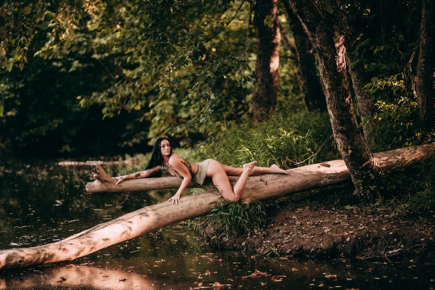 Woman in swimsuit lying on fallen log over forest river surrounded by lush green trees.