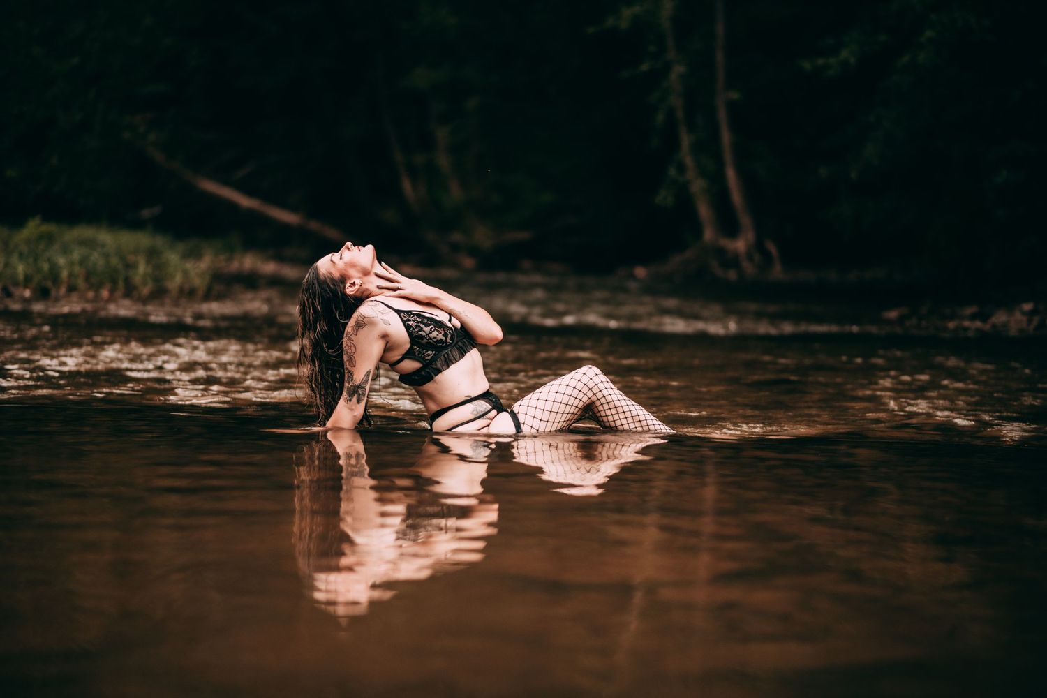 Woman in black lingerie and fishnet stockings posing gracefully in a shallow forest river.
