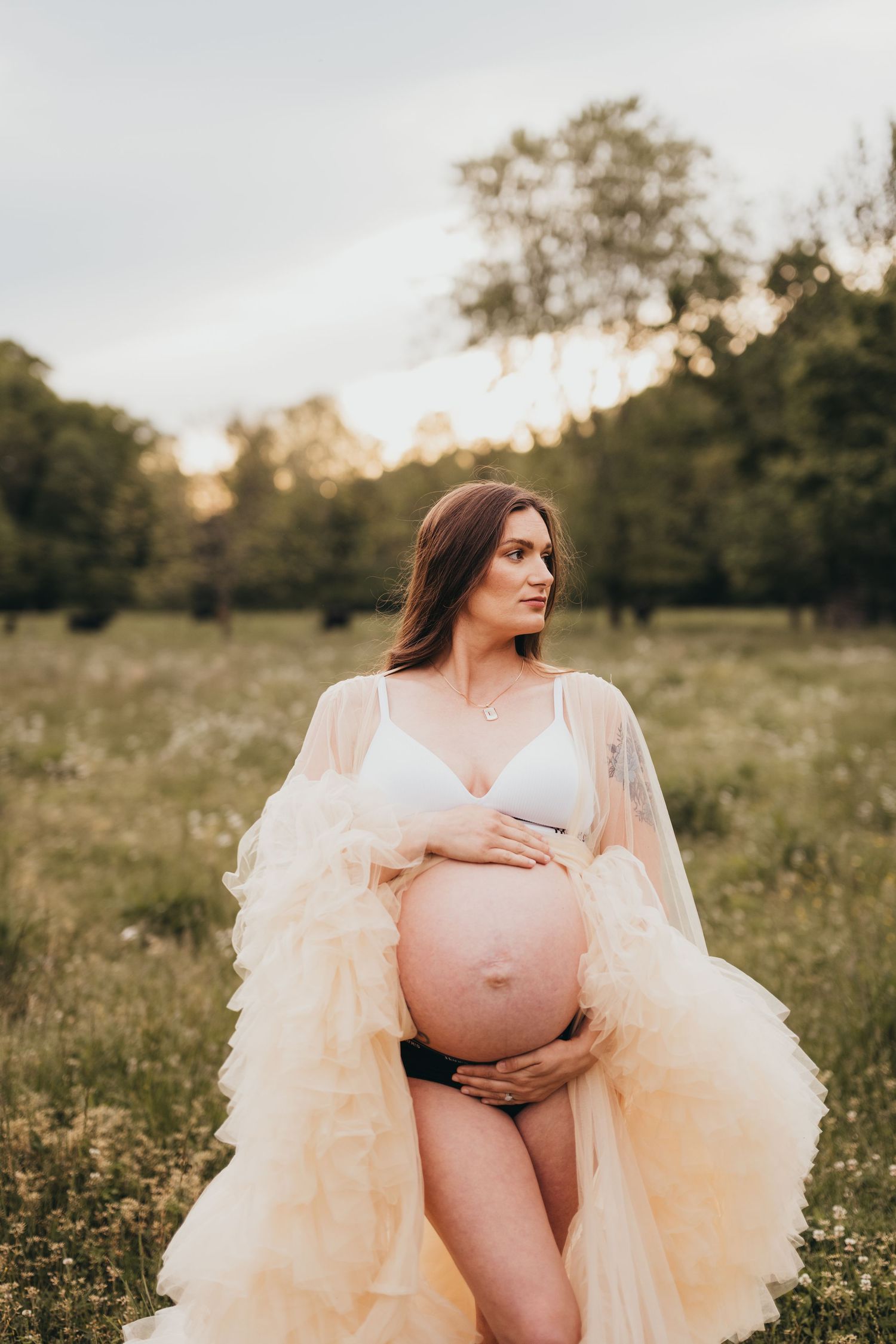 Dreamy maternity portrait in cream sheer robe amid wildflower field at golden hour.