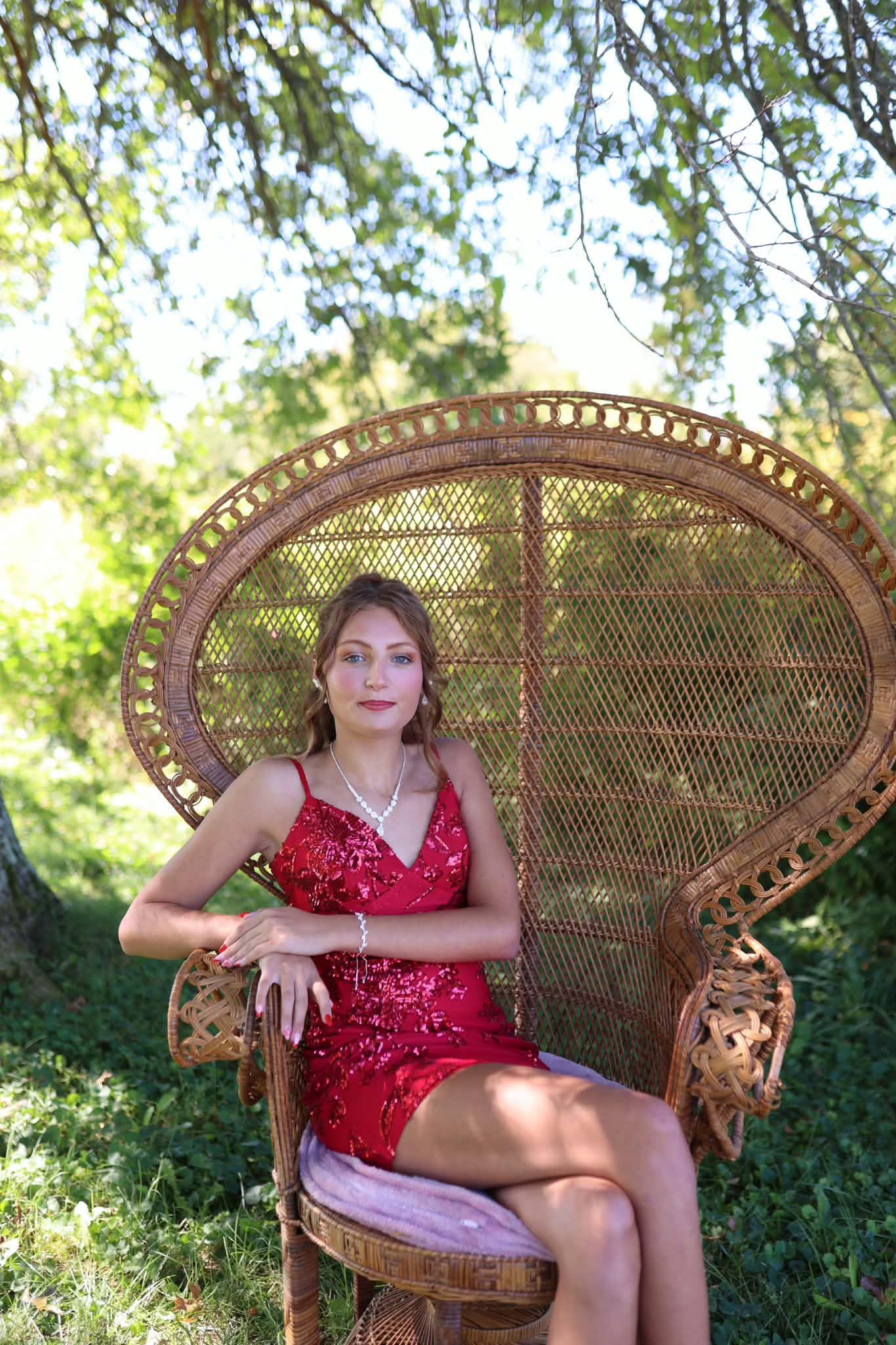 Young woman in red sequin dress seated in ornate wicker peacock chair outdoors under green trees.