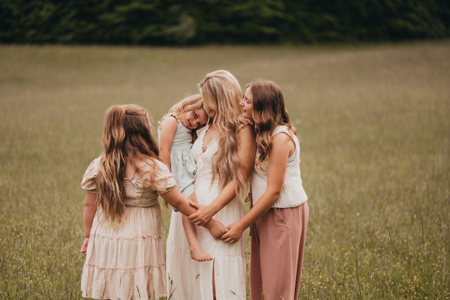A pregnant mother embraced by three daughters in a green meadow, wearing soft boho dresses in a tender family portrait.