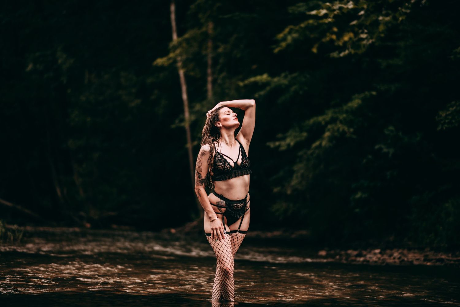 Woman in black lingerie and fishnet stockings standing in a forest river with lush green trees in background.