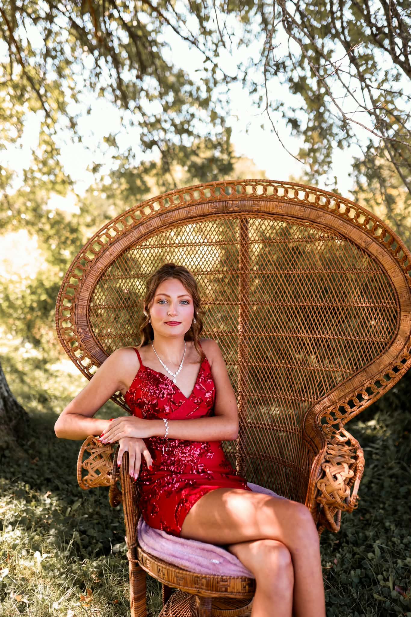 Young woman in red sequin dress seated in ornate wicker peacock chair outdoors among trees in golden light.