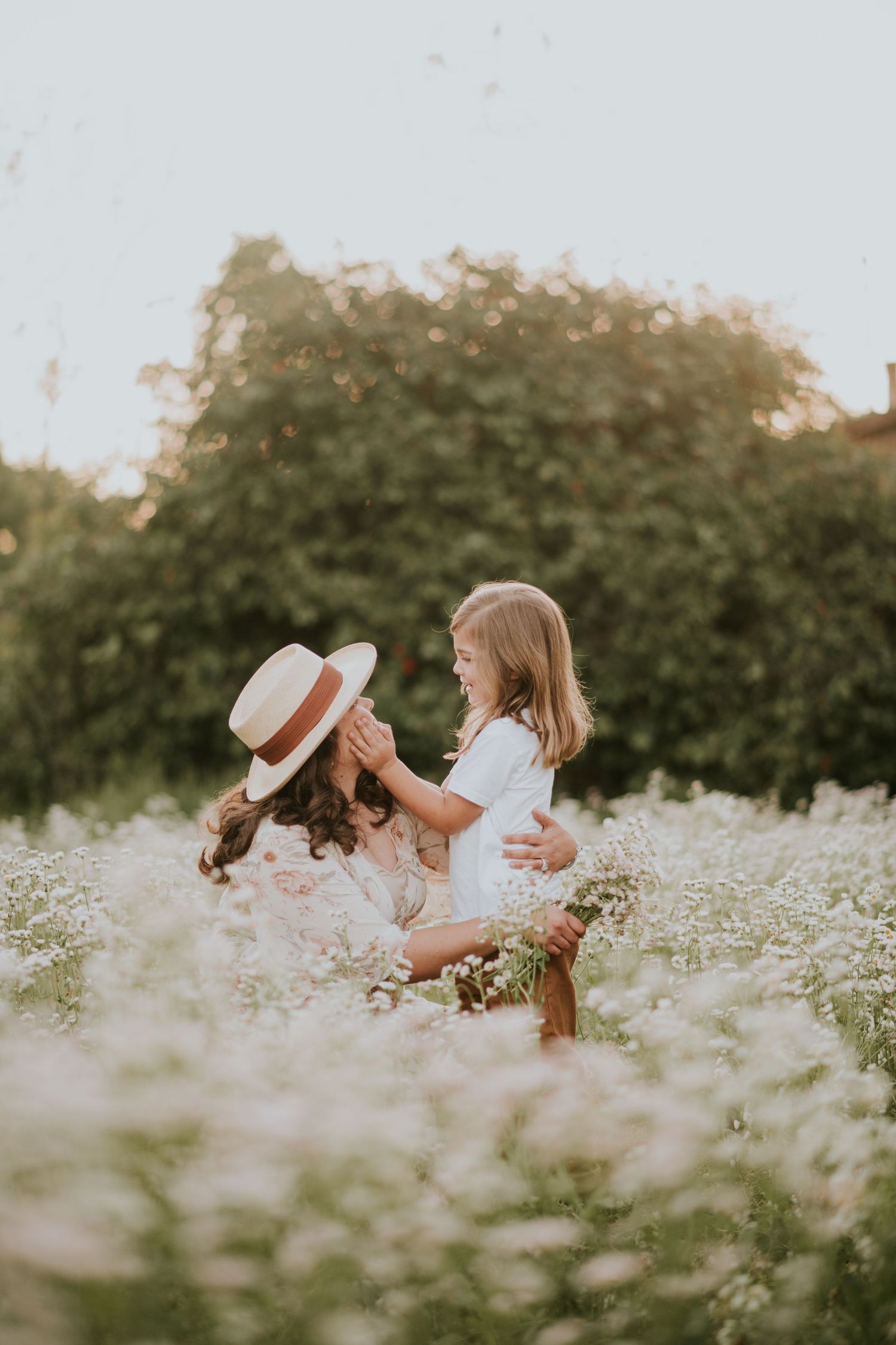A mother and daughter share a joyful moment in a blooming wildflower field, with the girl touching her mom's straw hat.