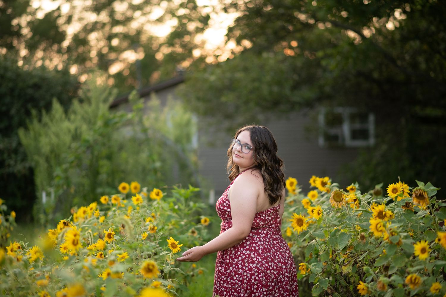 Woman in red floral dress standing among sunflowers in a garden at golden hour.